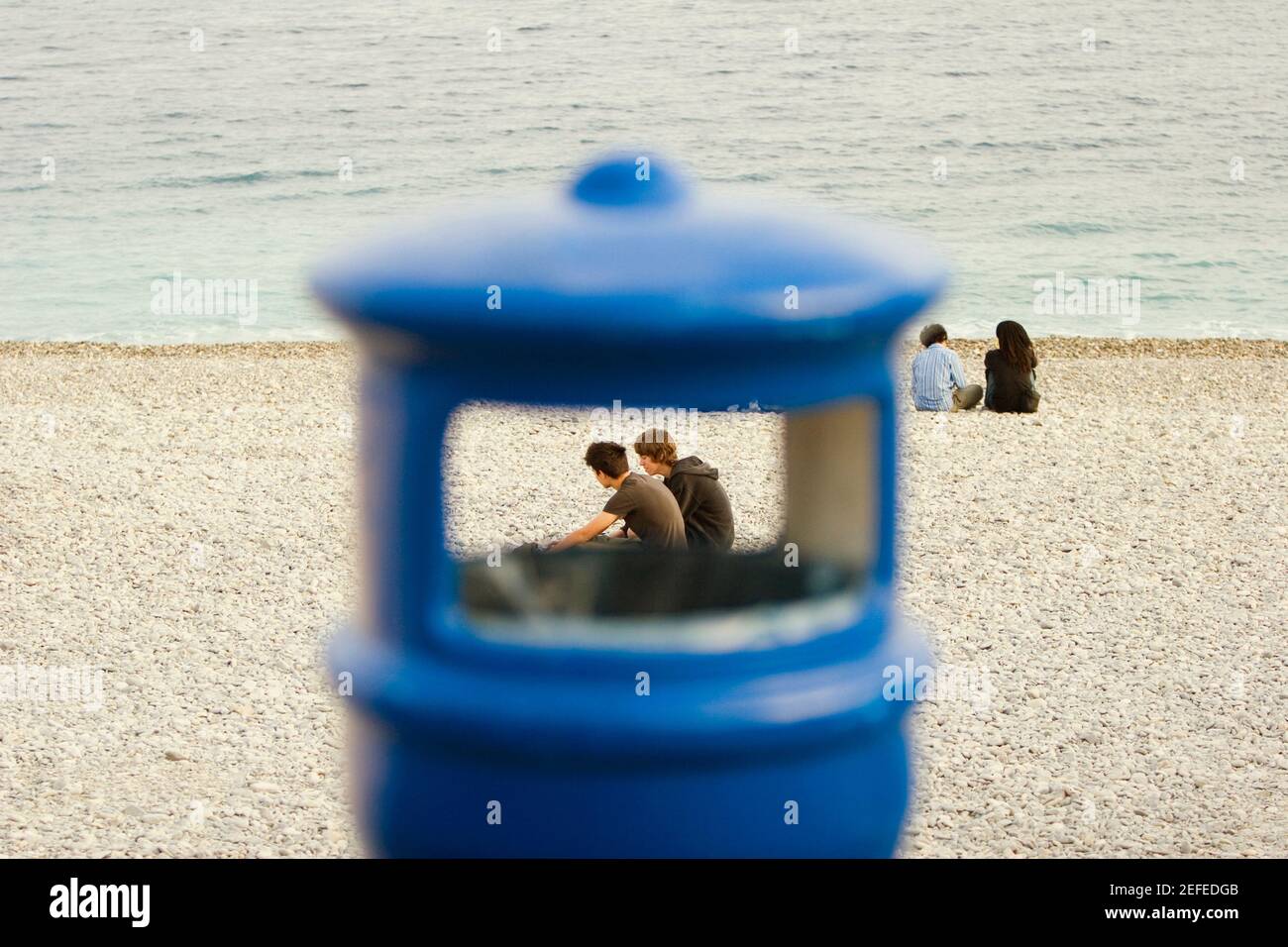 Couple sitting on the beach viewed through a garbage bin, Baie Des Anges, Nice, Provence Alpes Cote DÅ½Azur, France Stock Photo