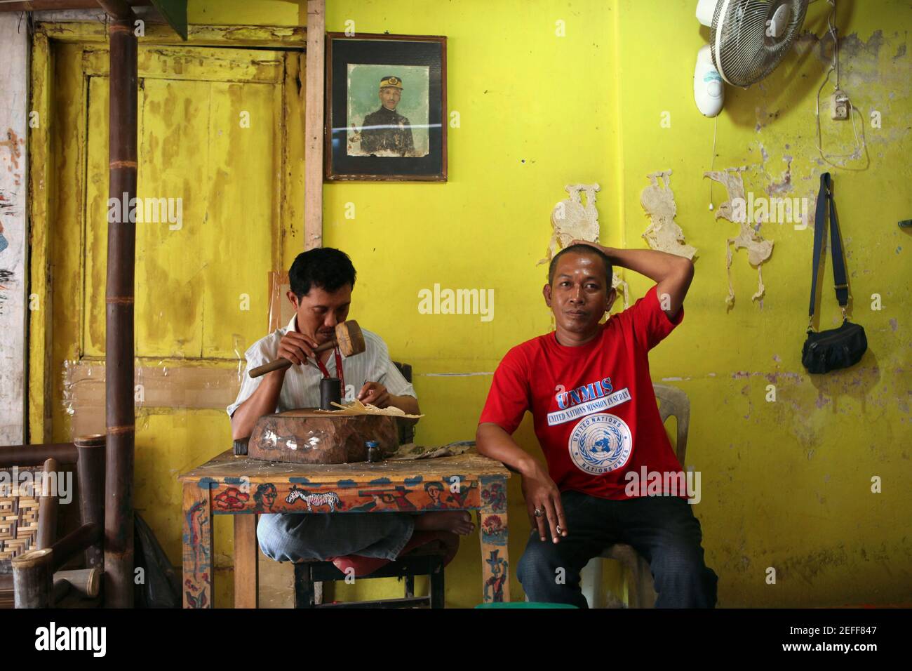 Craftsman makes a leather puppet for wayang kulit theatre in the Classic Wayang crafts shop in Yogyakarta, Central Java, Indonesia. Crafts shop manager Dodo wearing a blue shirt is pictured at the right. The portrait of the founder of the crafts shop is seen on the yellow wall. Kanjeng Raden Tumenggung Djojodiningrat, the great grandfather of the current owner of the crafts shop, was the court puppet master of the Sultan Hamengkubuwono IX of Yogyakarta. This is because the Classic Wayang is situated right in the Yogyakarta Keraton, the area of the Sultan palace.  Traditional puppet shadow thea Stock Photo