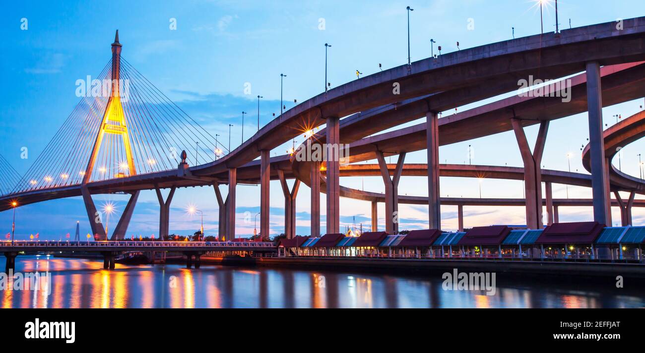 Picturesque underneath view of highway interchange and suspension bridges at dusk, glowing light reflection on a river. Bhumibol Bridge, Thailand. Stock Photo