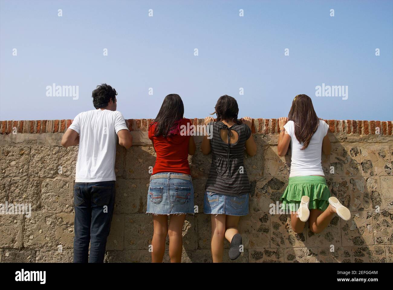 Rear view of three young women and a young man looking over a stone wall, Morro Castle, Old San Juan, San Juan, Puerto Rico Stock Photo