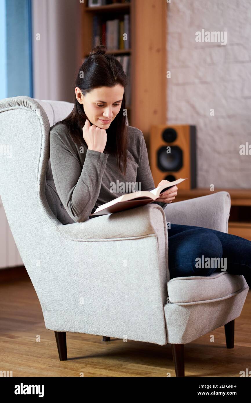 young woman sits on an armchair and reads a book Stock Photo