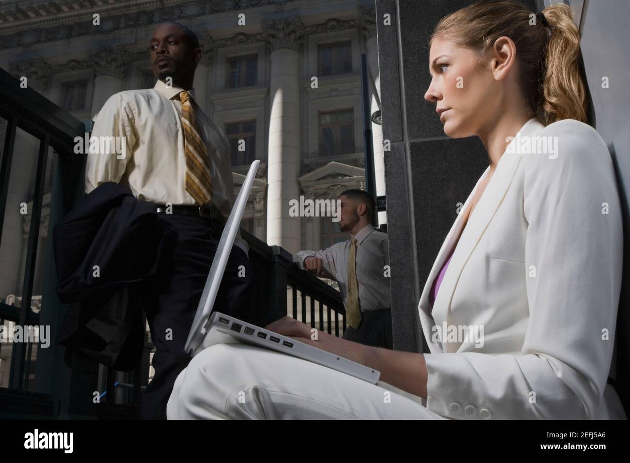 Side profile of a businesswoman using a laptop with two businessmen in the background Stock Photo
