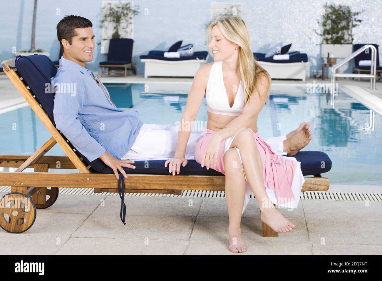 Young woman and a mid adult man sitting on a deck chair at the poolside ...