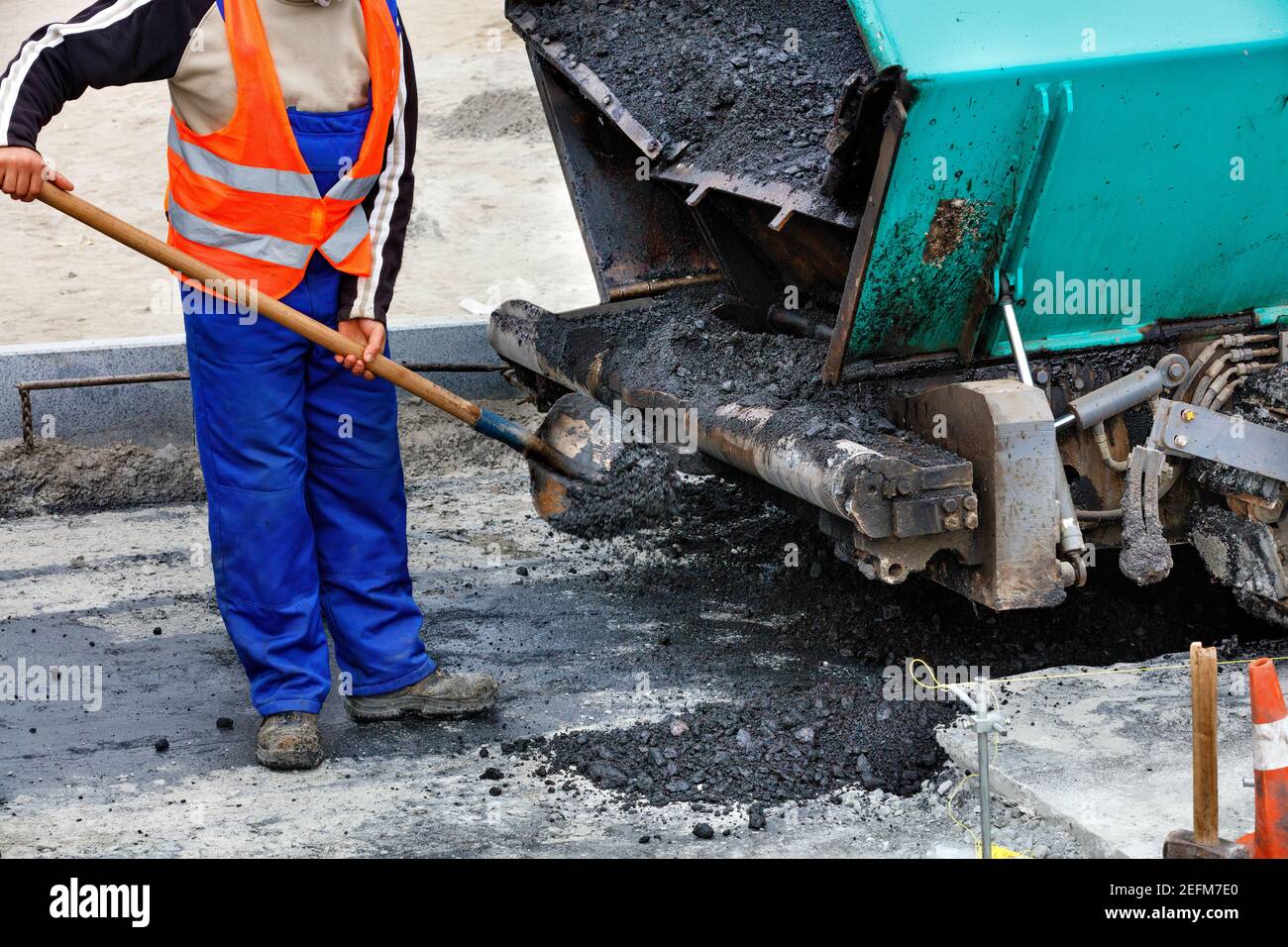 A worker in a blue overalls and an orange jacket cleans an asphalt paver with a shovel. Selective focus. Stock Photo