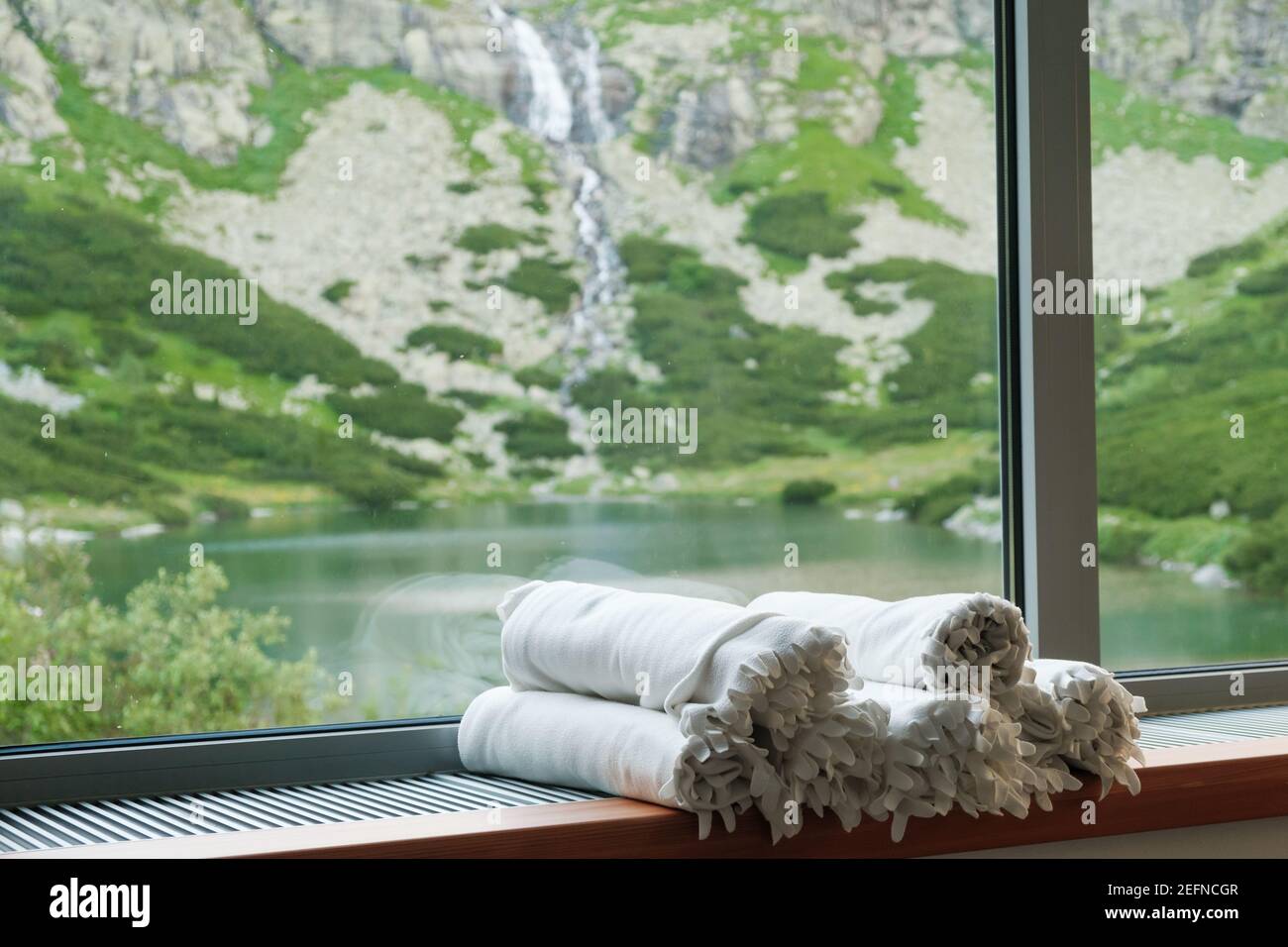 White blankets laying on the windowsill in front attractive view on the lake, waterfall and mountains. SPA and wellness concept. Stock Photo