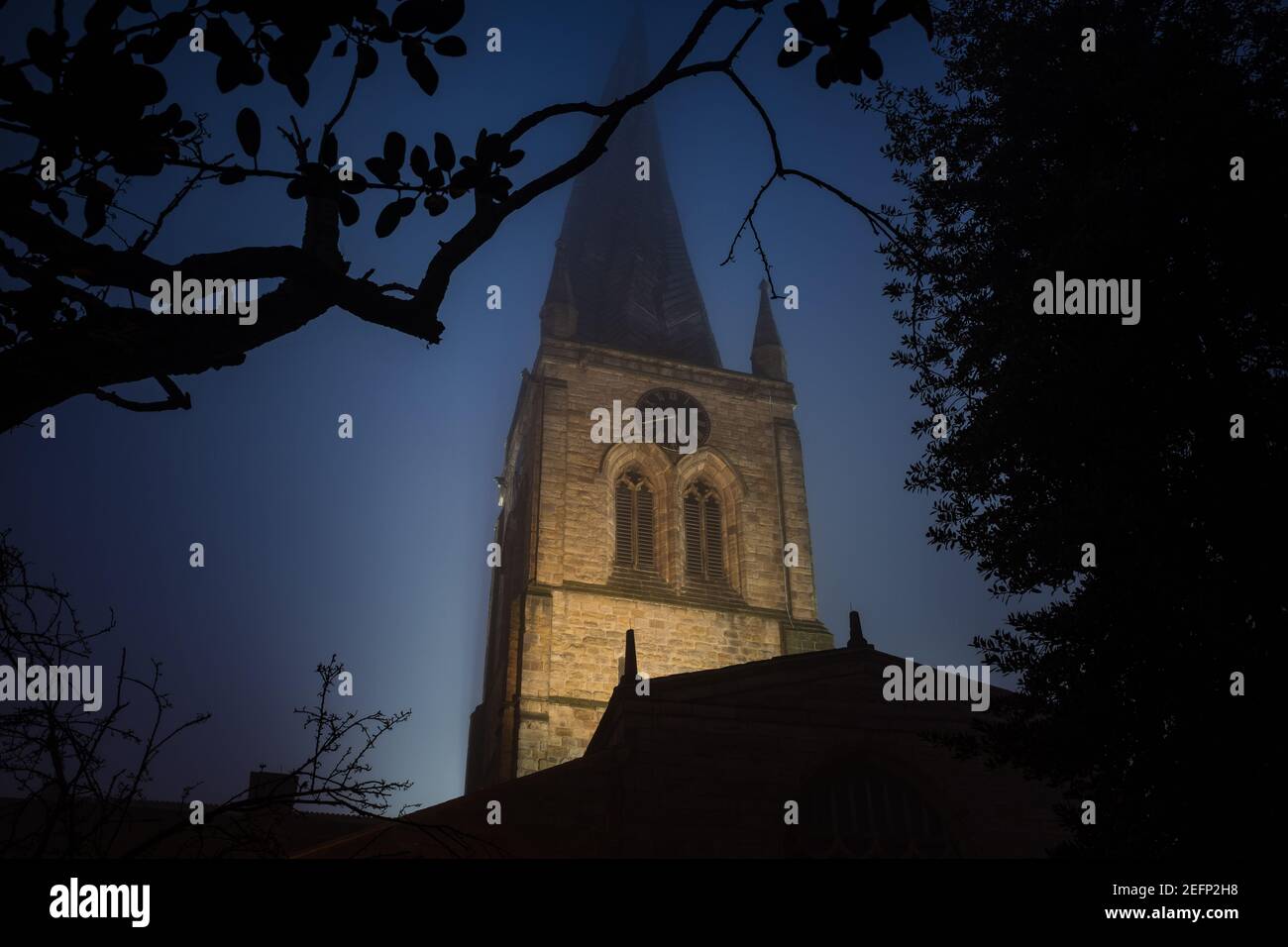 The Chesterfield Crooked Spire of St Marys church lit up illuminated during evening blue hour at night with fog and silhouette of trees Derbyshire Stock Photo