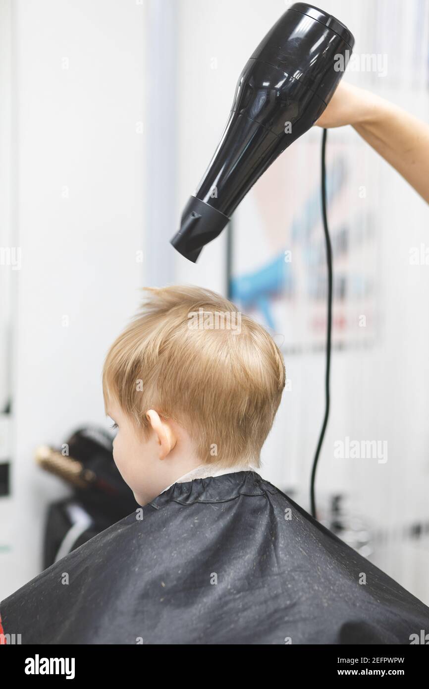 Hairdresser barbershop in beauty salon dries hair with large black hairdryer after cutting haircut for child, boy, caucasian, blond. Vertical shot. Stock Photo