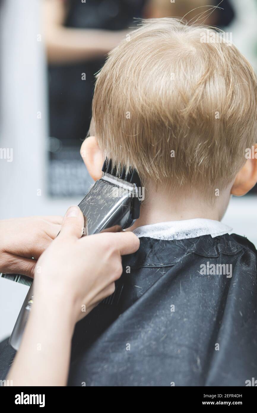 Hairdresser in barbershop machine clipper trimmer cuts hair child boy. Lifestyle close-up. Concept of beauty, hygiene and. Back view. Vertical shot of Stock Photo