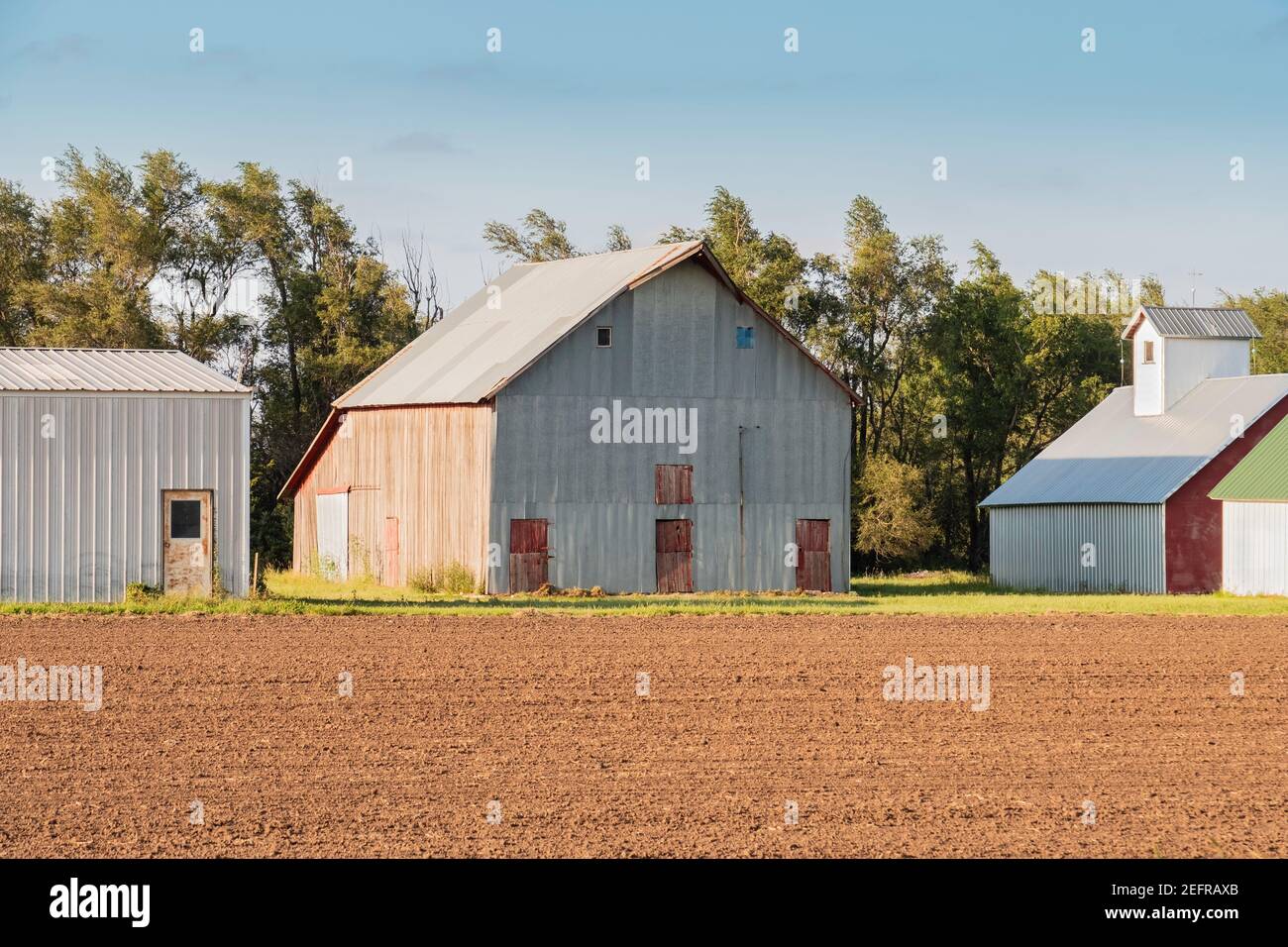 A farmyard with a metal and wood barn, garage and outbuilding. Plowed field. Kansas, USA. Stock Photo
