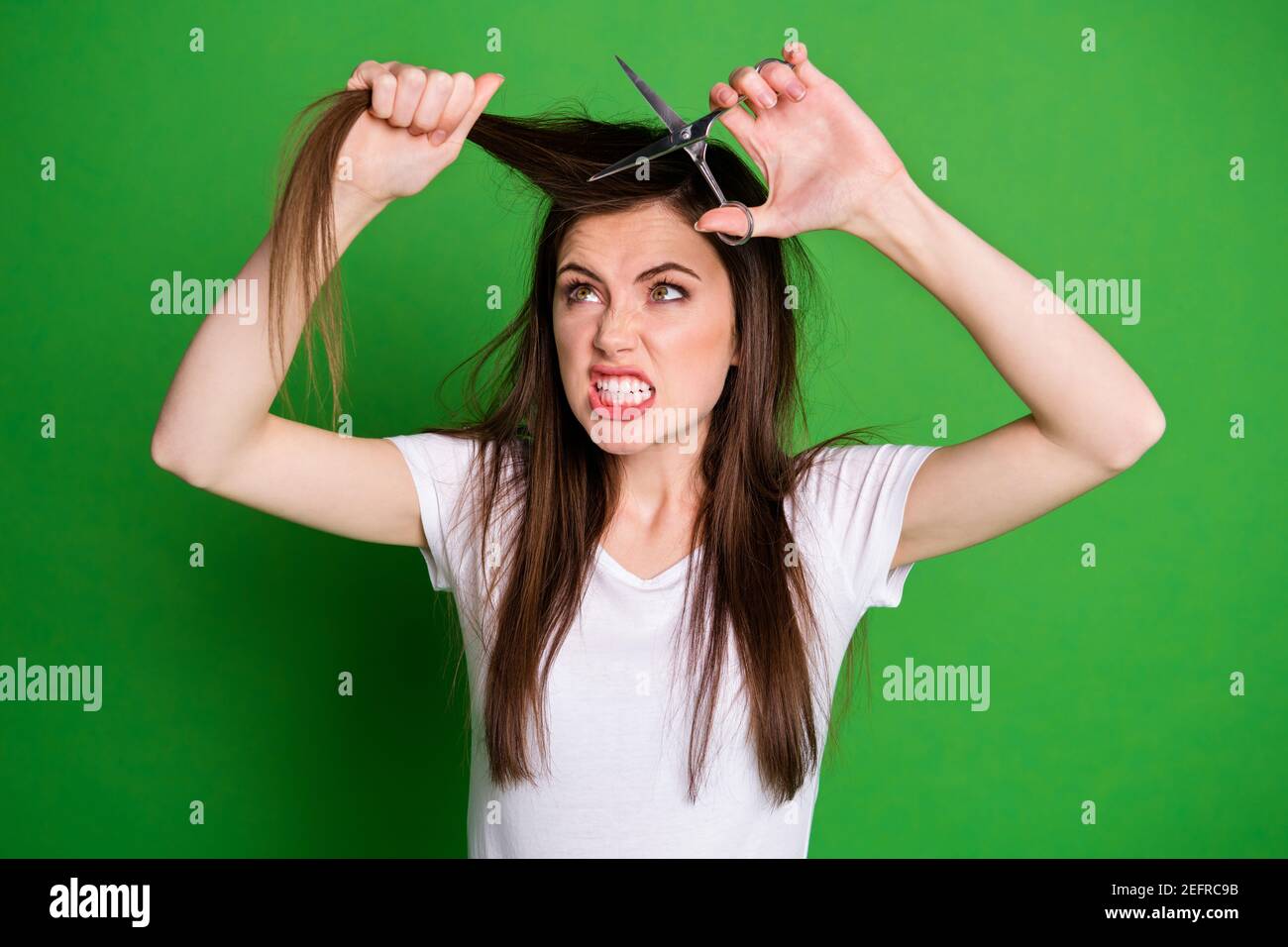 Photo portrait of angry woman cutting hair with scissors isolated on vivid green colored background Stock Photo