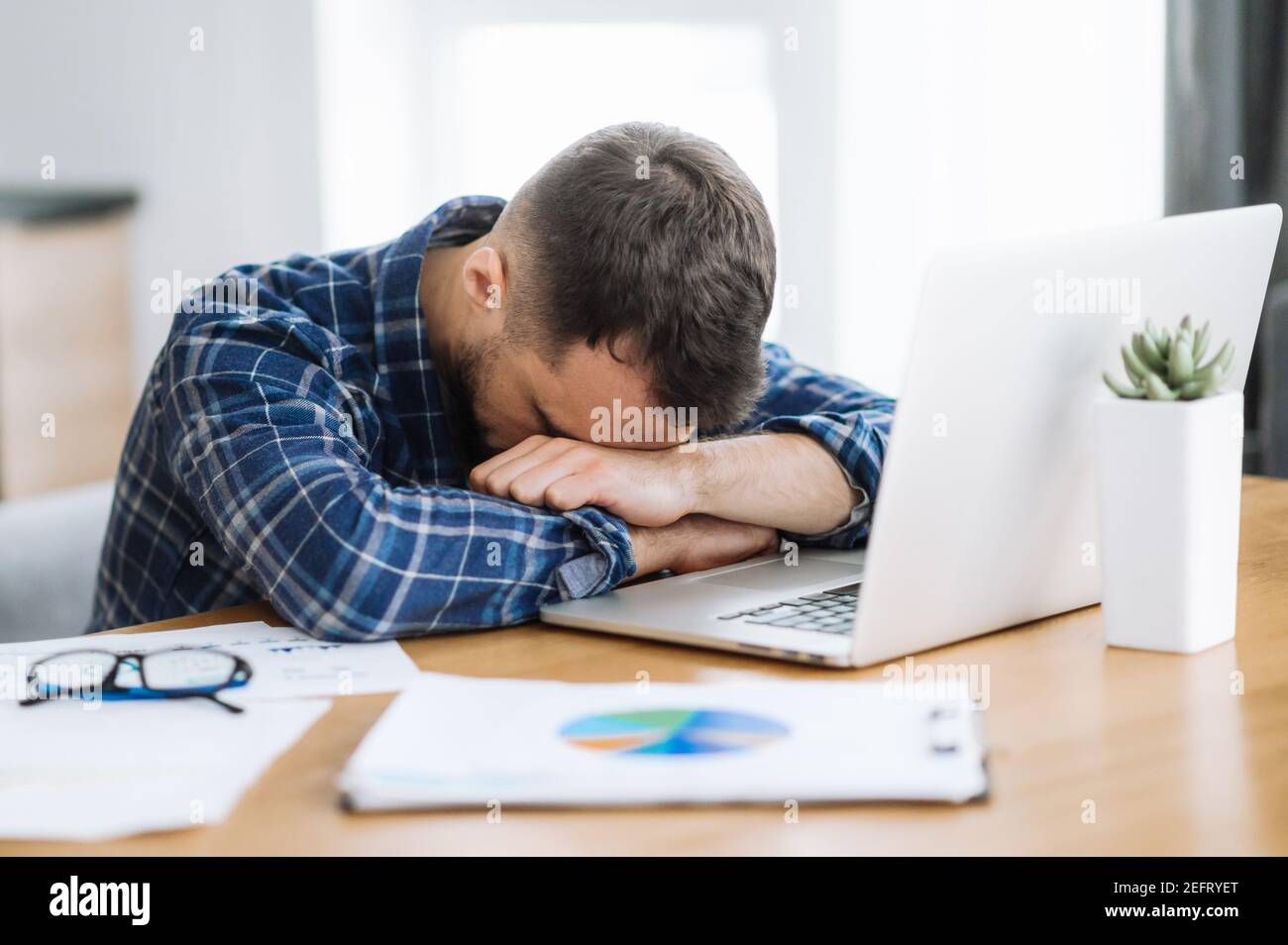 Tired freelancer man is fall asleep at the workplace. Exhausted busy guy working overtime, sits at the desk at home, need a rest, overwork concept. Work from home Stock Photo