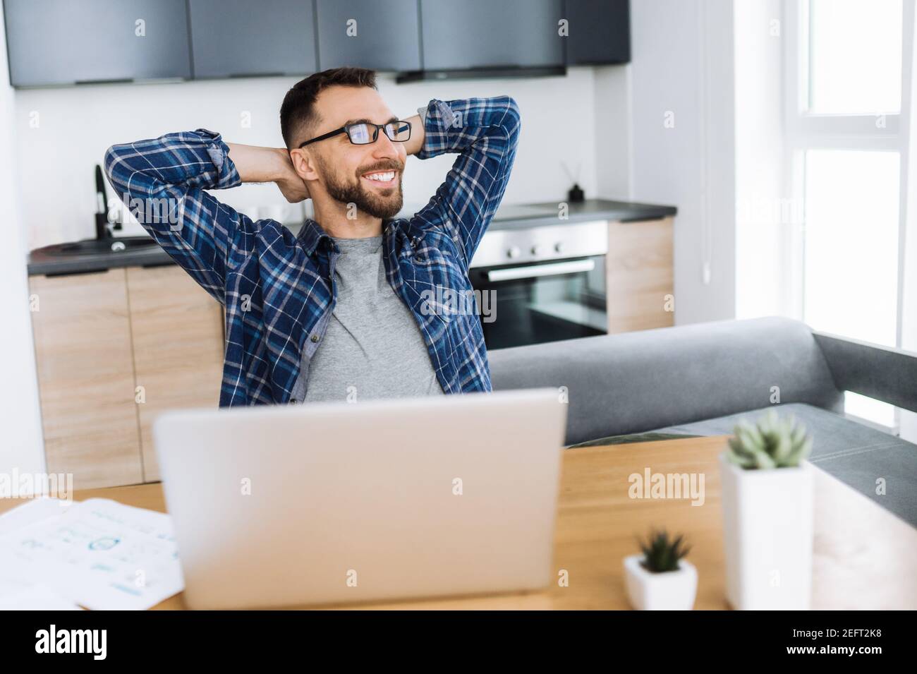 Positive freelancer guy relaxing at the workplace and smiling. Confident caucasian male entrepreneur sitting at home office, taking break from distant working and dreaming about vacation Stock Photo