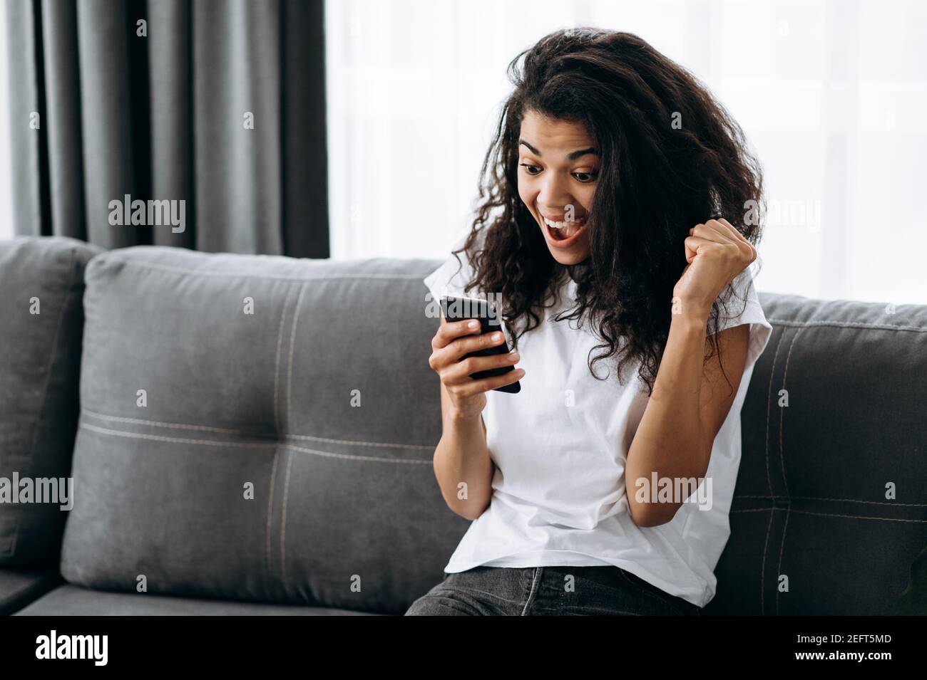 Joyful young lady sitting on the couch, using smartphone. Excited beautiful woman look at the screen, winning at the phone game or reading good news, pass exams or get a dream job Stock Photo
