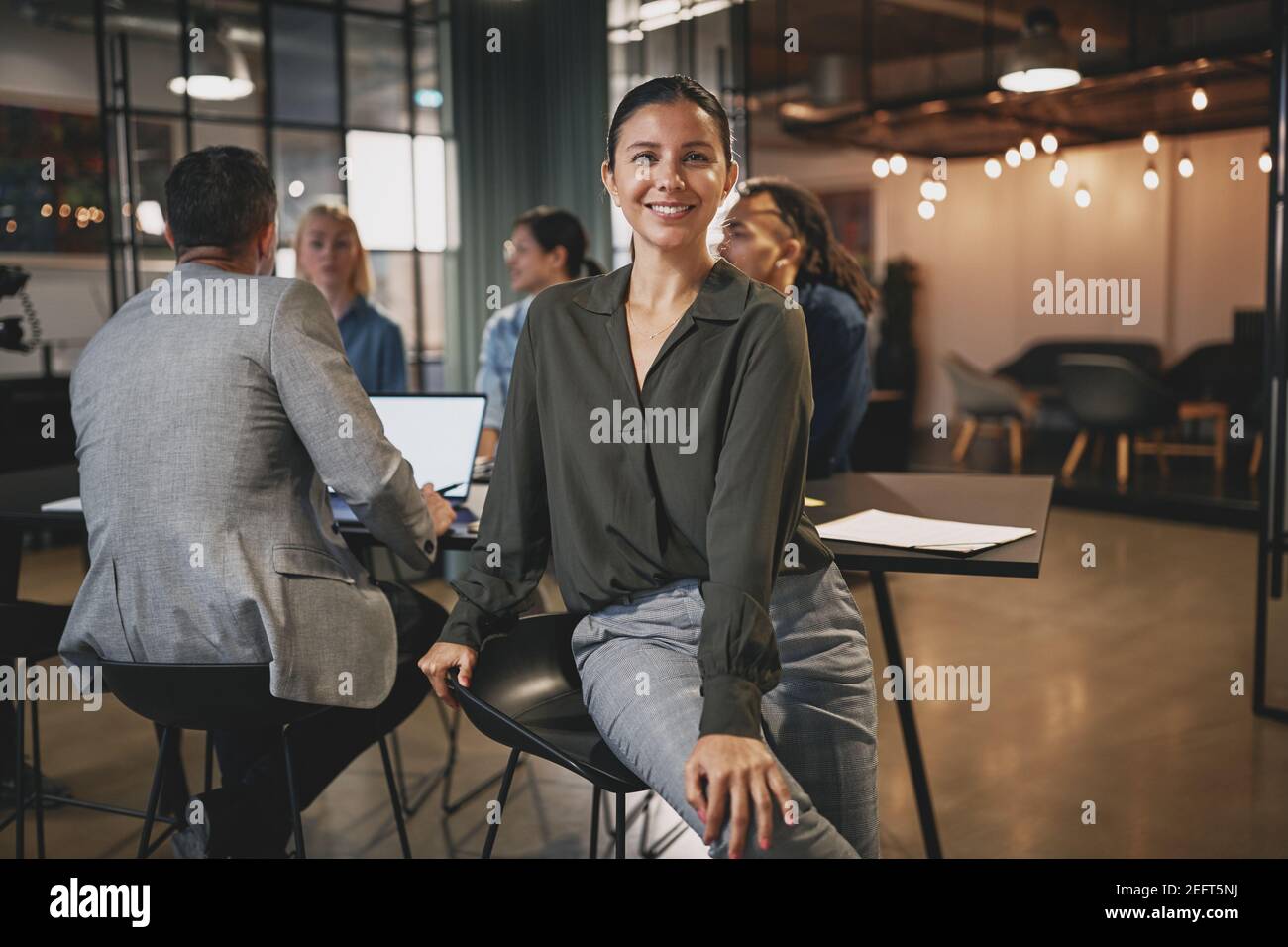 Young businesswoman smiling while sitting in an office with coworkers talking together at a table in the background Stock Photo