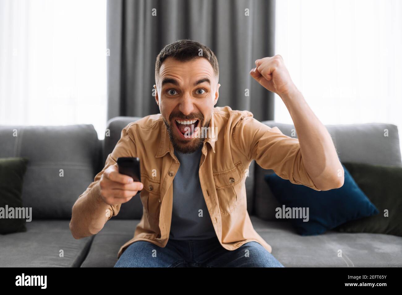 Excited joyful man sitting on the sofa, watching on tv sport channel. Cheerful emotional guy support the team, screaming, rejoices of successful game, football fan Stock Photo