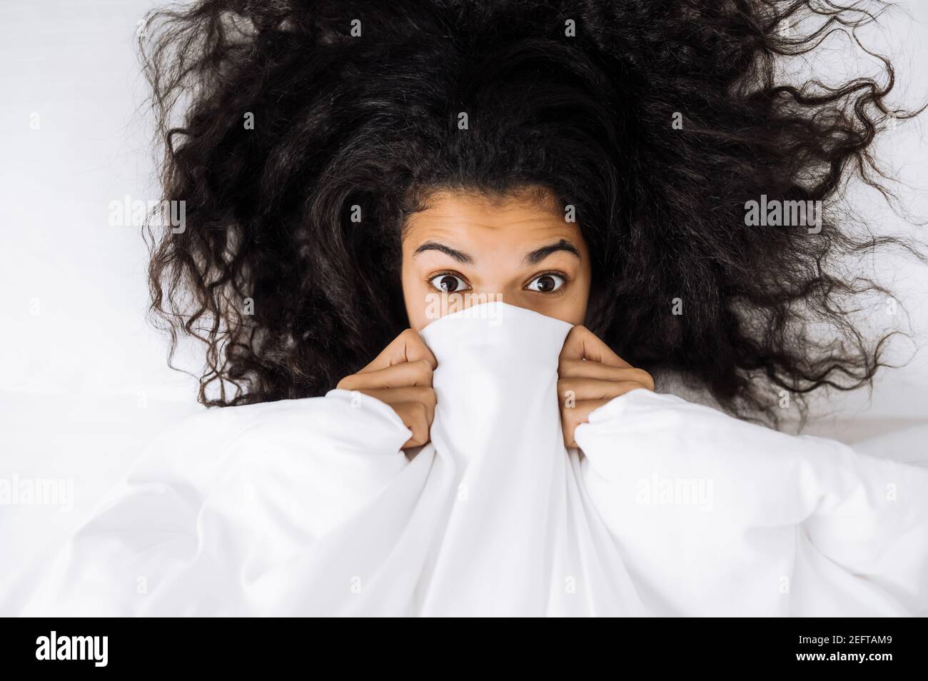 Portrait of adorable young lady with curly hair, under white blanket.  Attractive girl looking at the camera, waking up after good night sleep,  chilling at home in the morning Stock Photo -