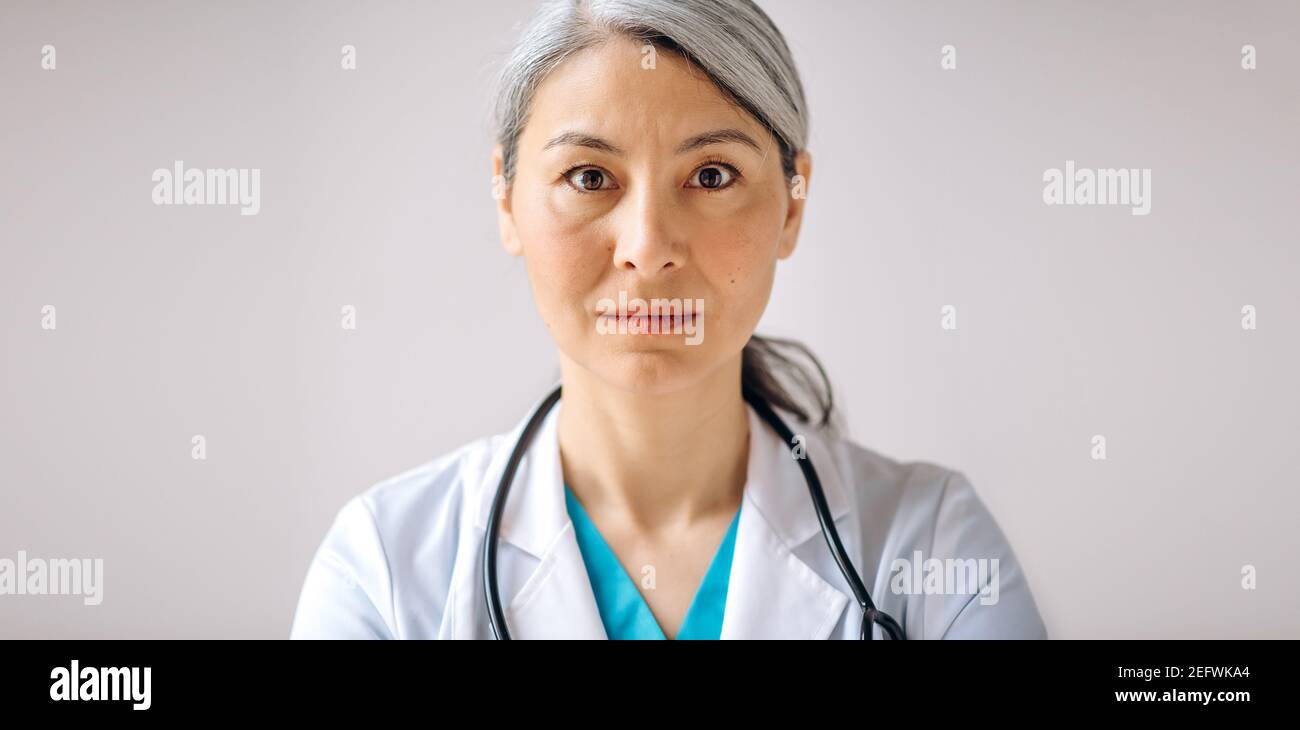 Panoramic portrait of asian gray-haired female doctor, general practitioner or pediatrician in medical uniform and stethoscope, standing against isolated background and looking directly at the camera Stock Photo