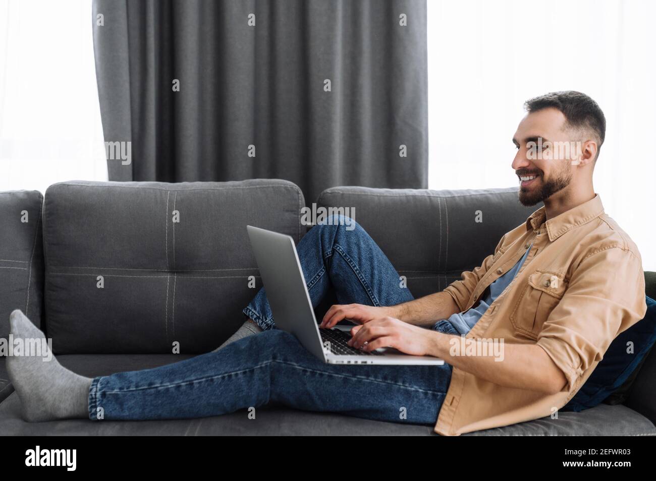 Caucasian male freelancer sits on the sofa, chilling at home. Bearded guy in casual wear use a laptop, watching films or funny videos, or working from home and smiling Stock Photo