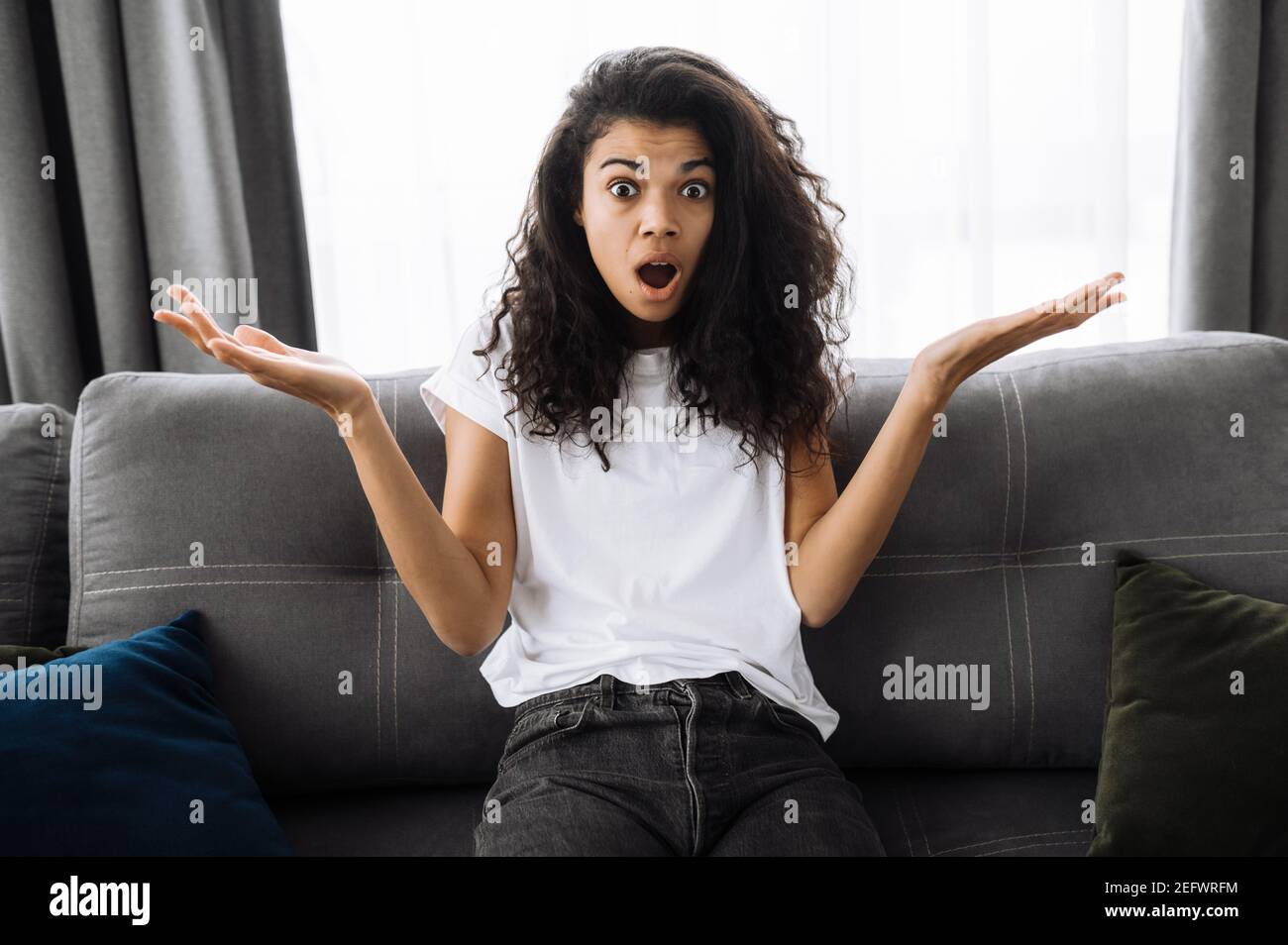 Portrait of surprised African American girl with curly hair. Shocked young woman in casual wear sits on the sofa at home, got unexpected news, shrugs up, and confused looks directly at the camera Stock Photo