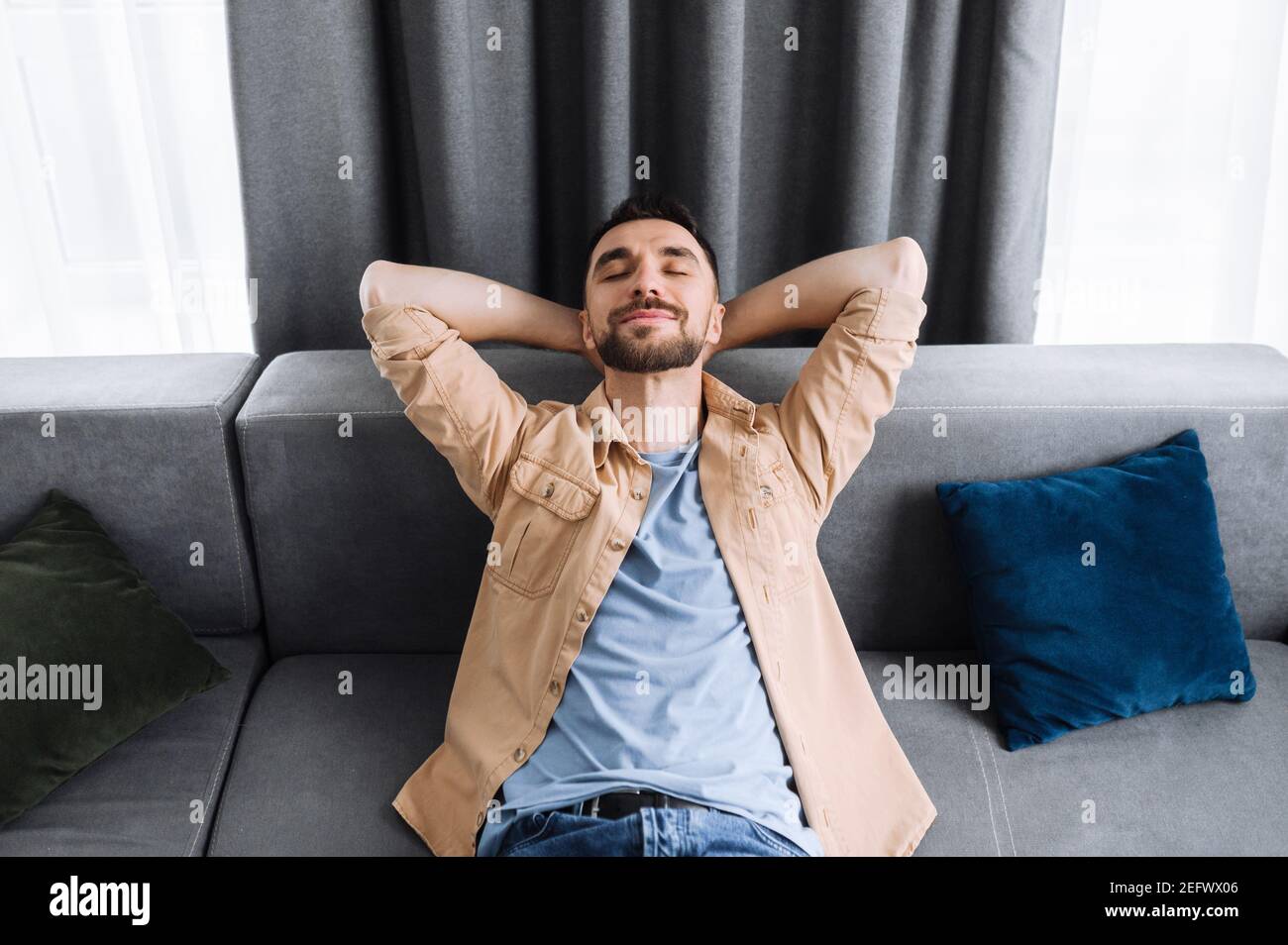 Attractive man is relaxing on the couch in living room at home and smiling. Caucasian stylish guy takes a break from online work, or have weekend, dreaming about vacations Stock Photo