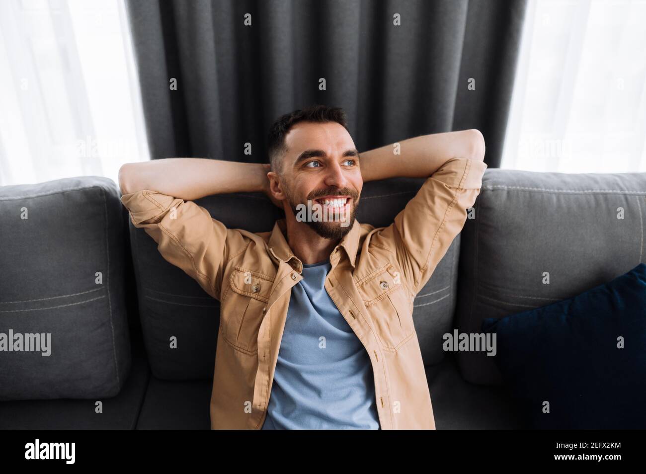 Portrait of a happy calm caucasian man sitting on the sofa, in the end of working day, resting from tense work, dreaming about vacations and smiling Stock Photo
