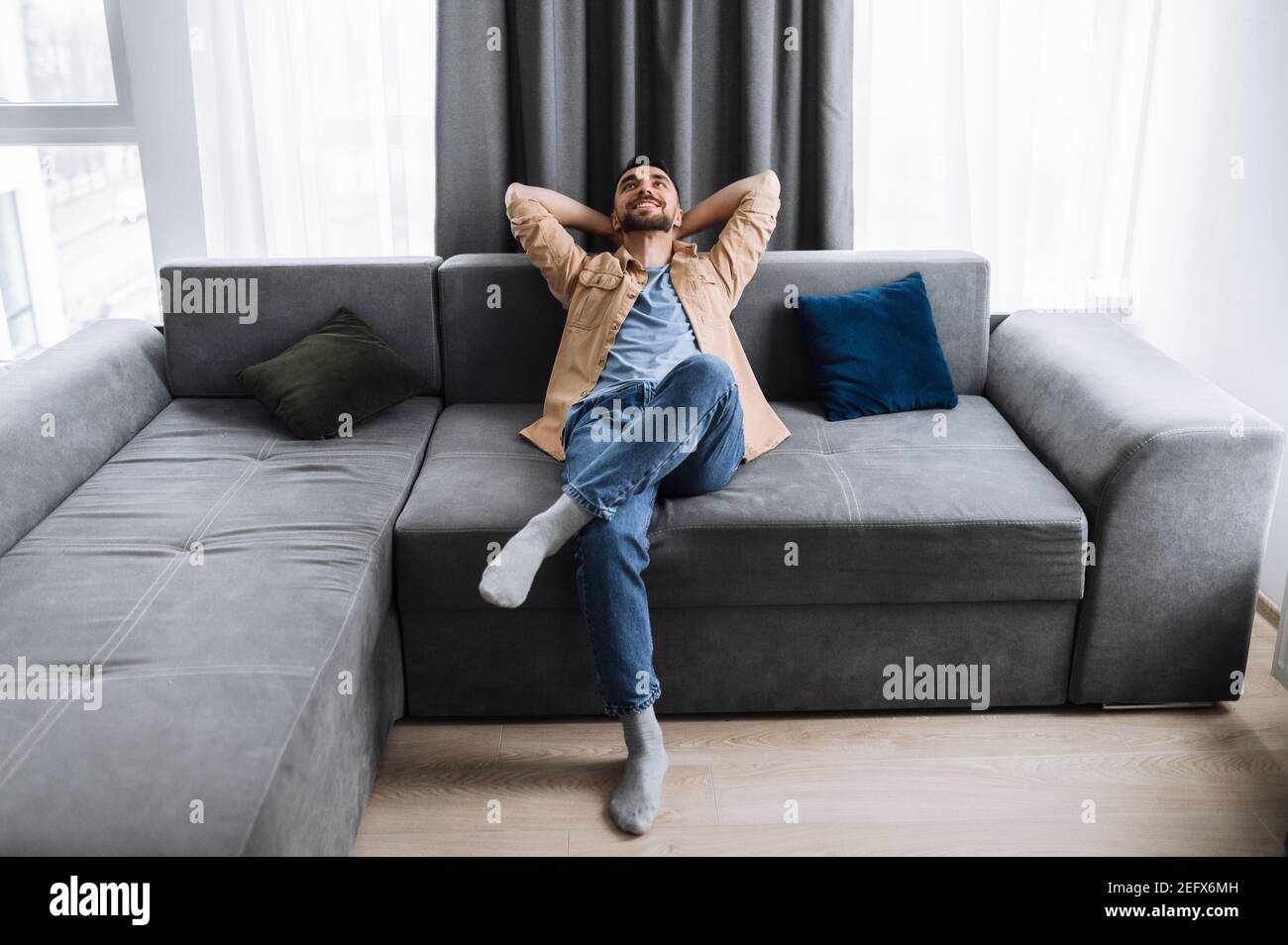 Satisfied handsome caucasian man is sitting on the couch in living roo, thinking about plans for weekends or vacation and smile. Young freelancer rest from online work Stock Photo