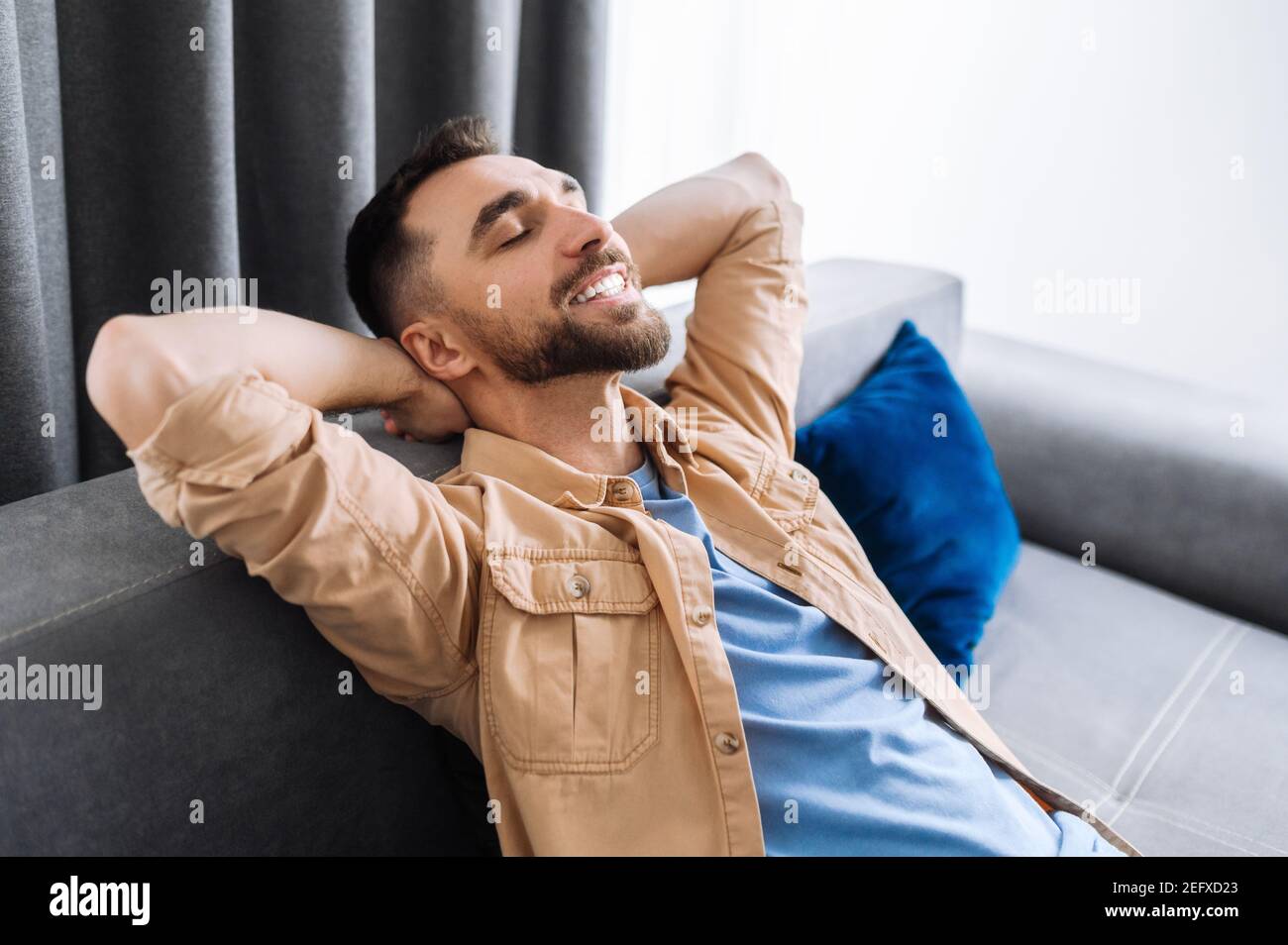 Attractive man is relaxing on the couch in living room at home and smiling. Caucasian stylish guy takes a break from online work, or have weekend, dreaming about vacations Stock Photo