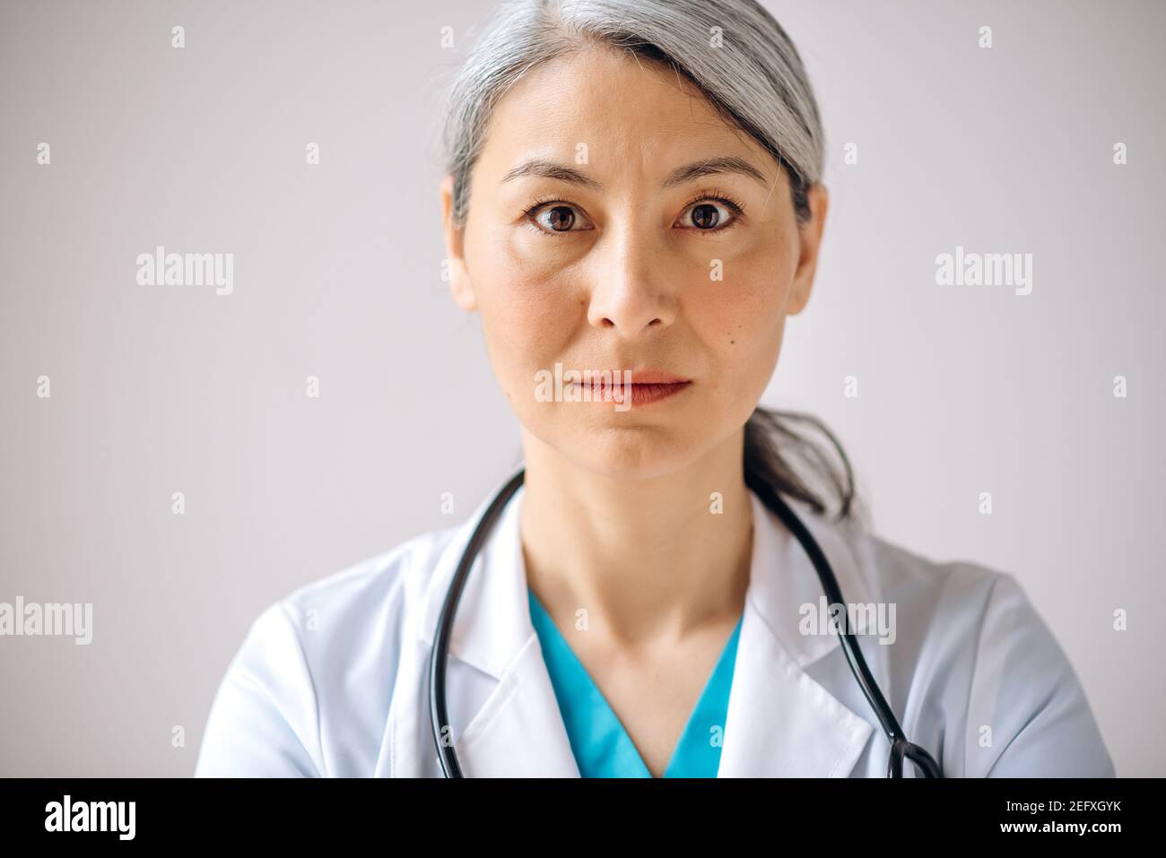 Close-up portrait of asian gray-haired female doctor, general practitioner or pediatrician in medical uniform and stethoscope, standing against isolated background and looking directly at the camera Stock Photo