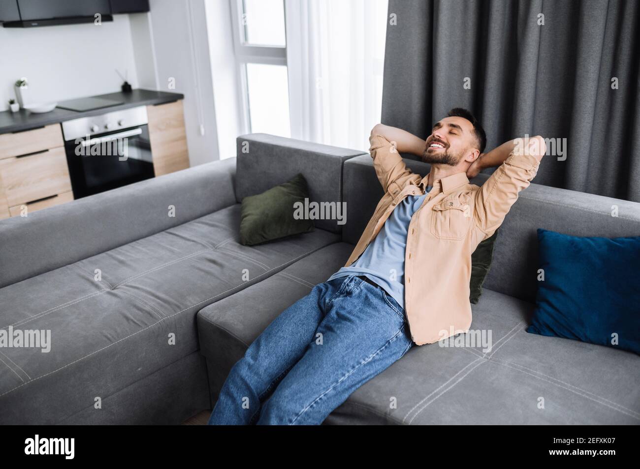 Satisfied calm handsome caucasian guy is sitting on the couch with closed eyes in living roo, thinking about plans for weekends or vacation and smile. Young freelancer rest from online work Stock Photo