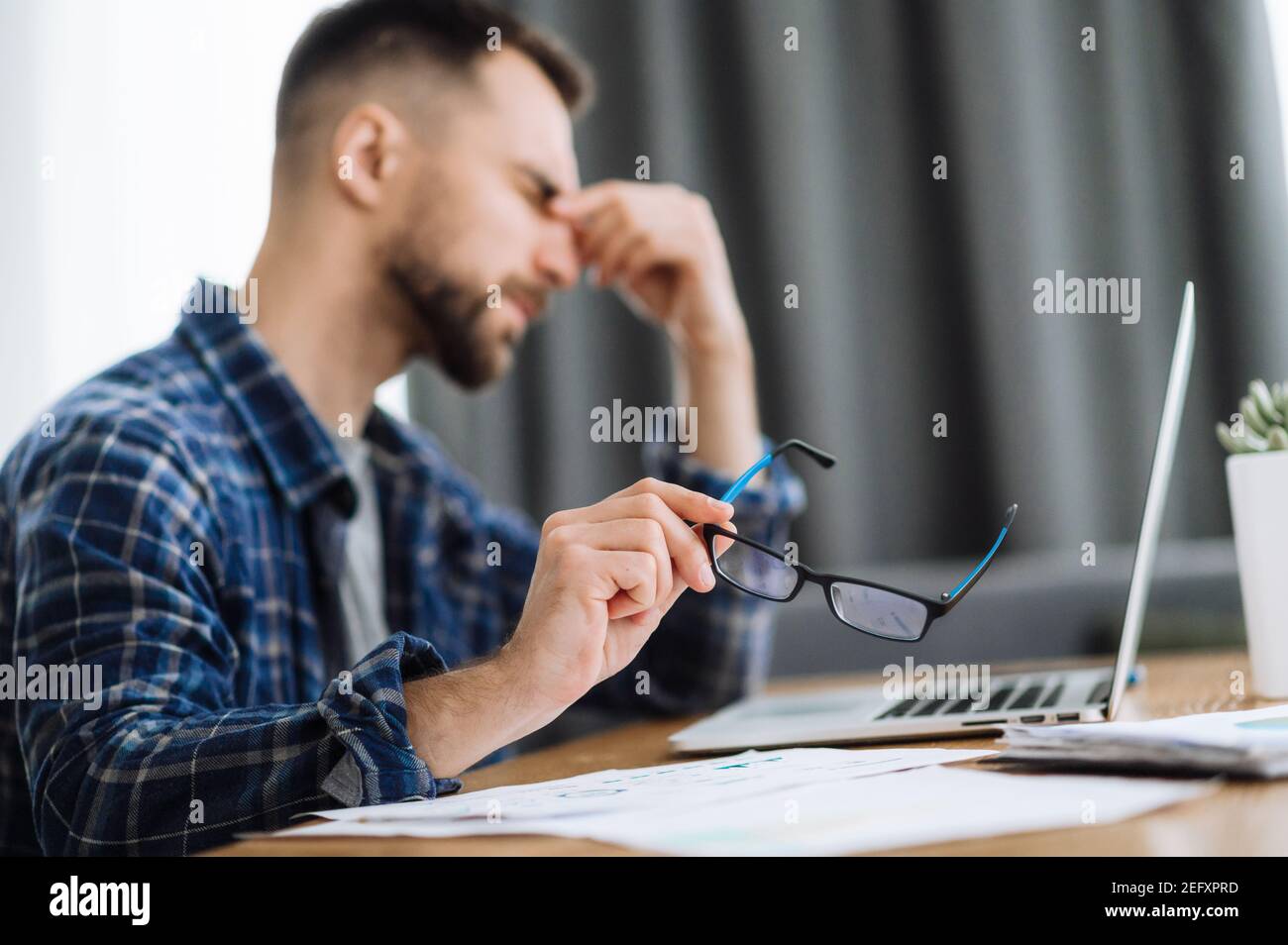 Tired male freelancer takes his glasses off and hold his hand on his face with with closed eyes, sitting at the work desk in home office, exhausted from work. He is rubbing the eyes, need a rest Stock Photo