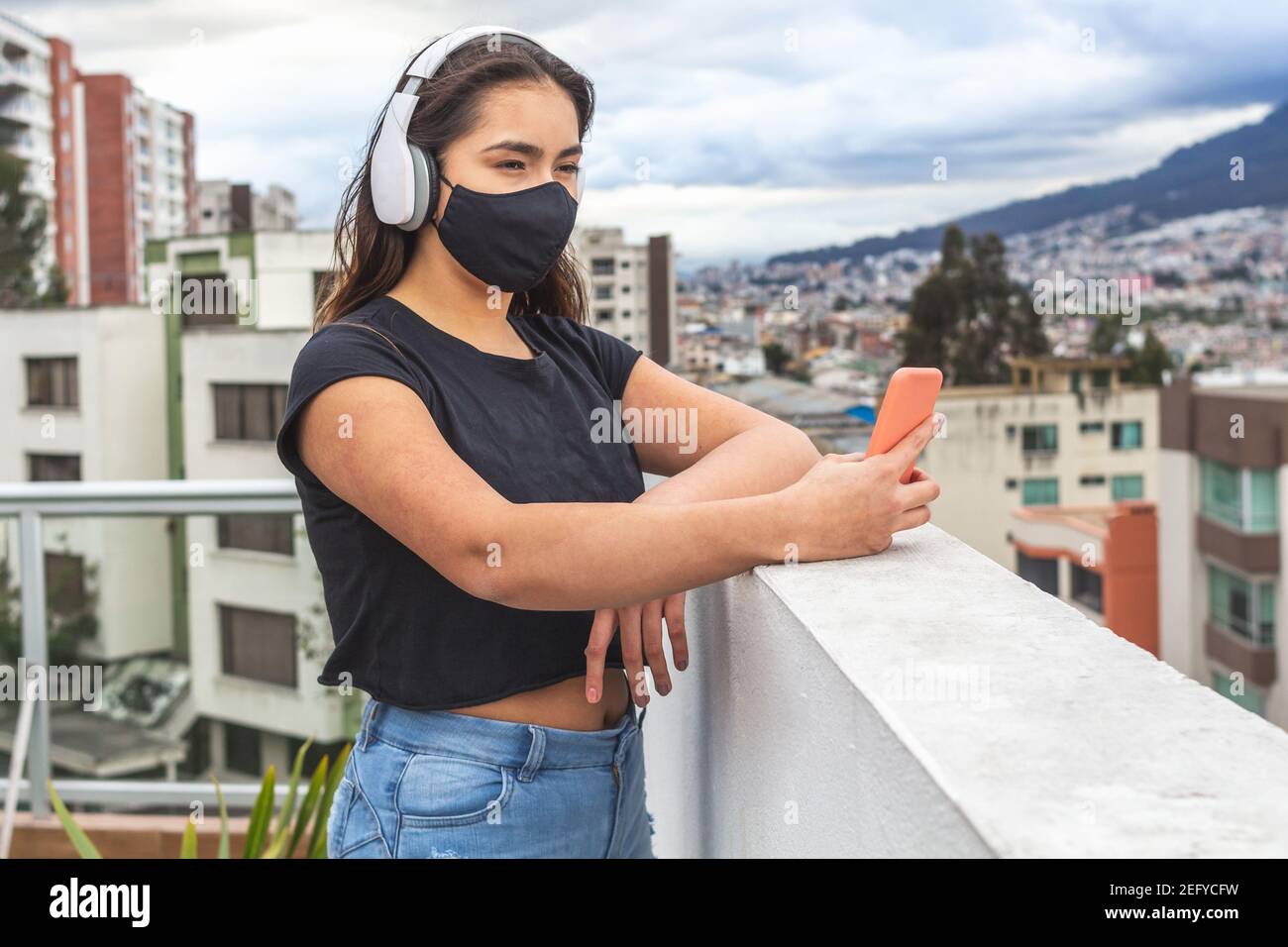 Teenage girl with a mask listening to music with wireless headphones and taking a selfie with her cell phone on a terrace or balcony outside of the ci Stock Photo
