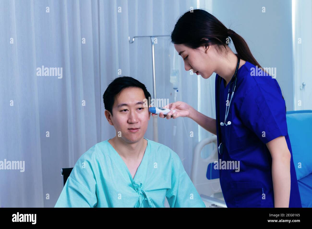 Young female medical worker with stethoscope examining ears of Asian male patient in uniform using tympanic thermometer for temperature in hospital Stock Photo