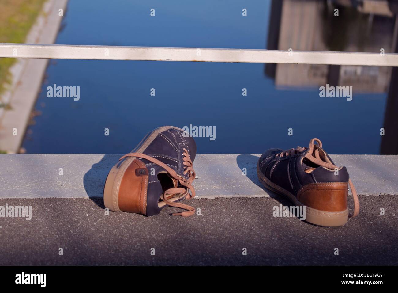 A pair of teenage dark sneakers with brown backs lying against a white railing on the edge of a bridge over the river below. The concept of suicide by Stock Photo