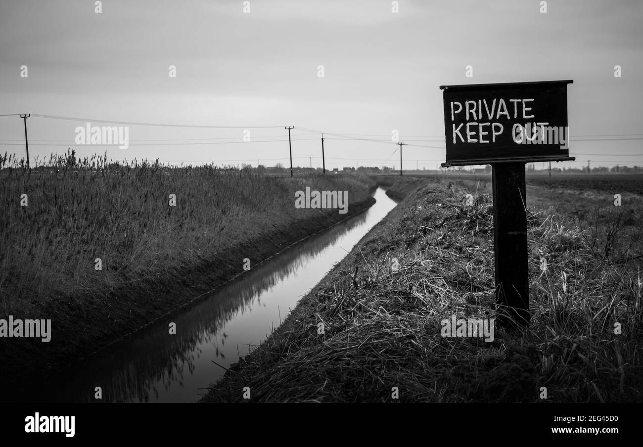 A Private Keep Out sign in an agricultural field in the Lincolnshire Fens next to a drainage dyke. Bleak and black & white Stock Photo