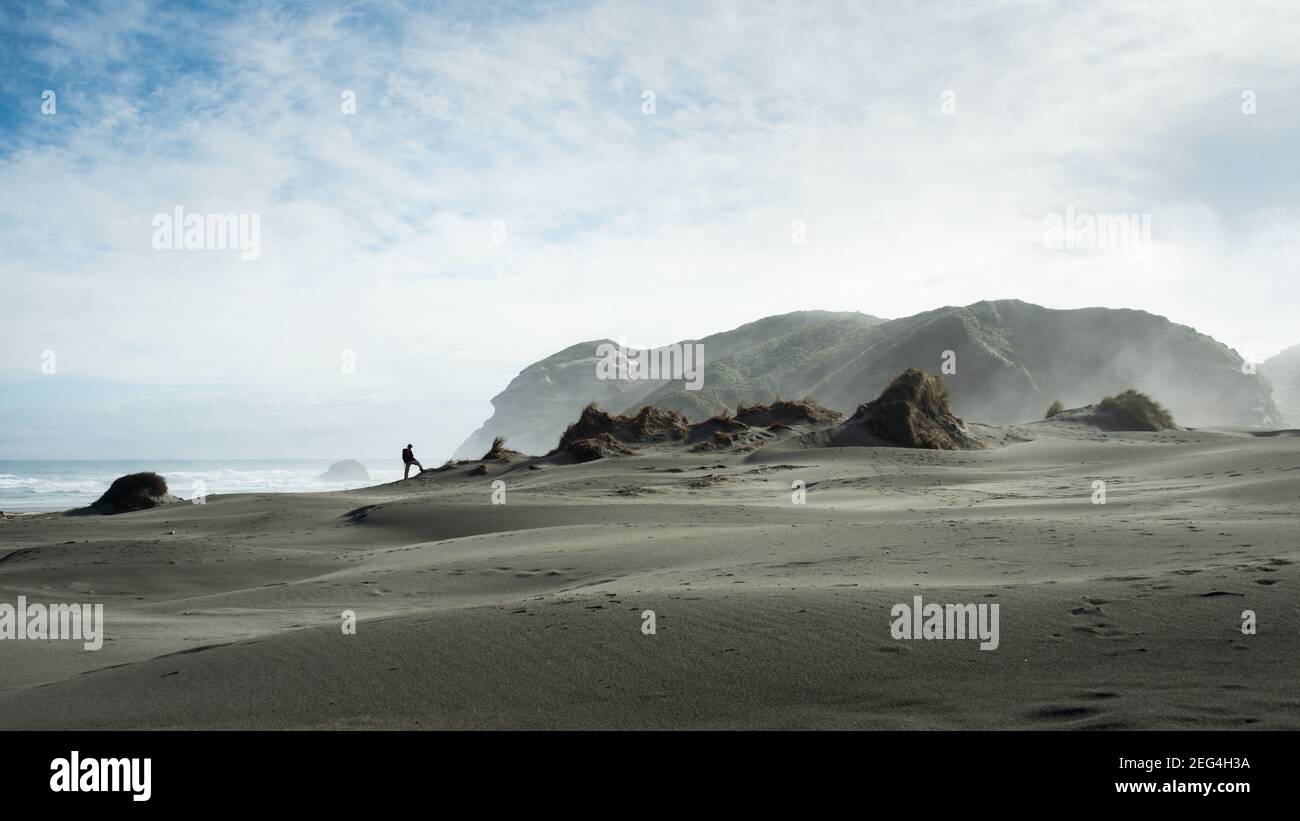 A backpacker standing on the sand dune with wind blowing the sands up in the air. Photo taken at Wharariki beach track, Golden Bay, South Island Stock Photo