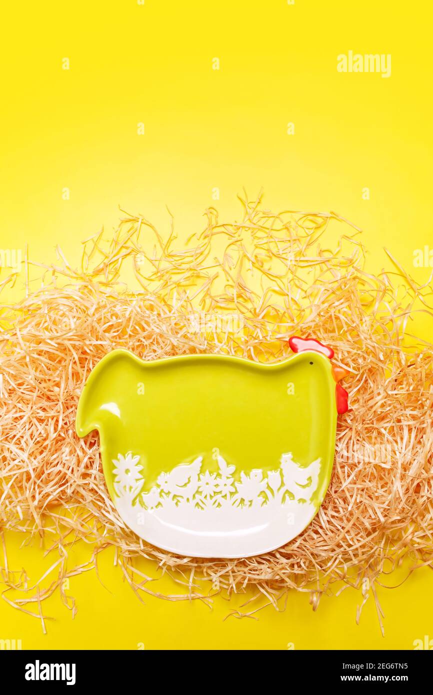 Easter frame background with green funny hen plate on hay over bright yellow color, flat lay