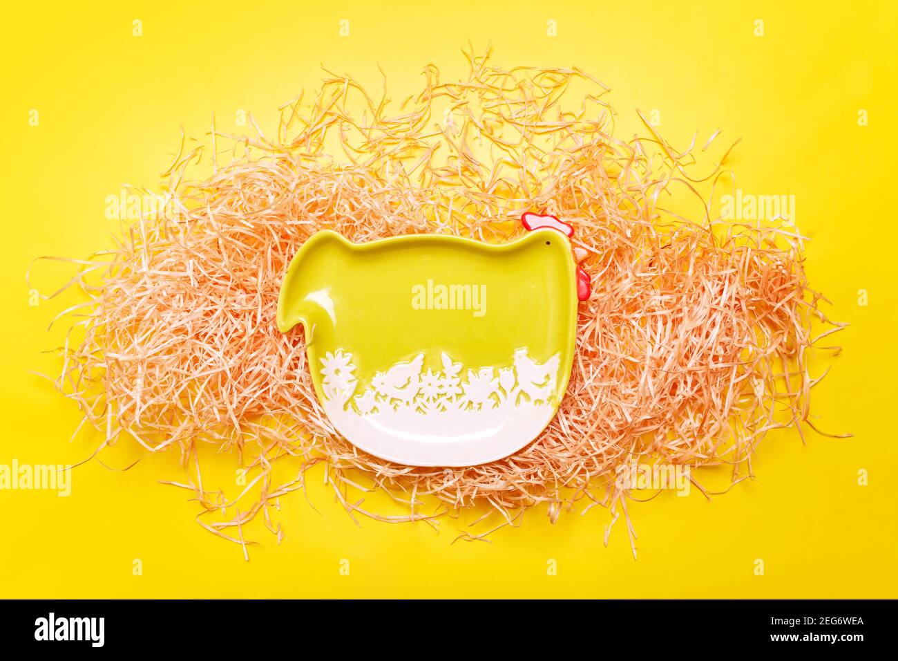 Easter frame background with green funny hen plate on hay over bright yellow color, flat lay