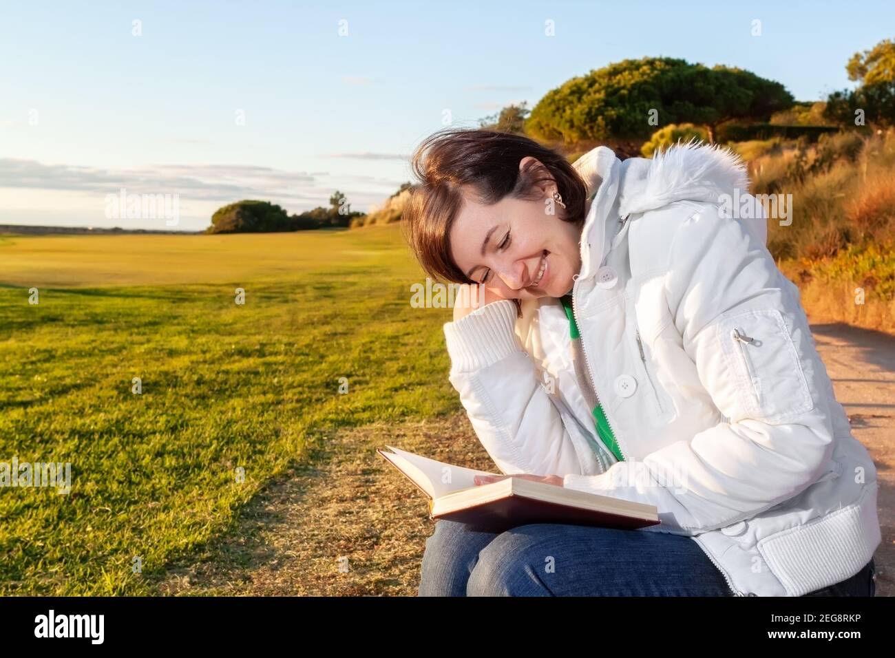 Dreamy photo of middle aged woman reading a book while sitting on a nice grassy area, during the sunset. Stock Photo