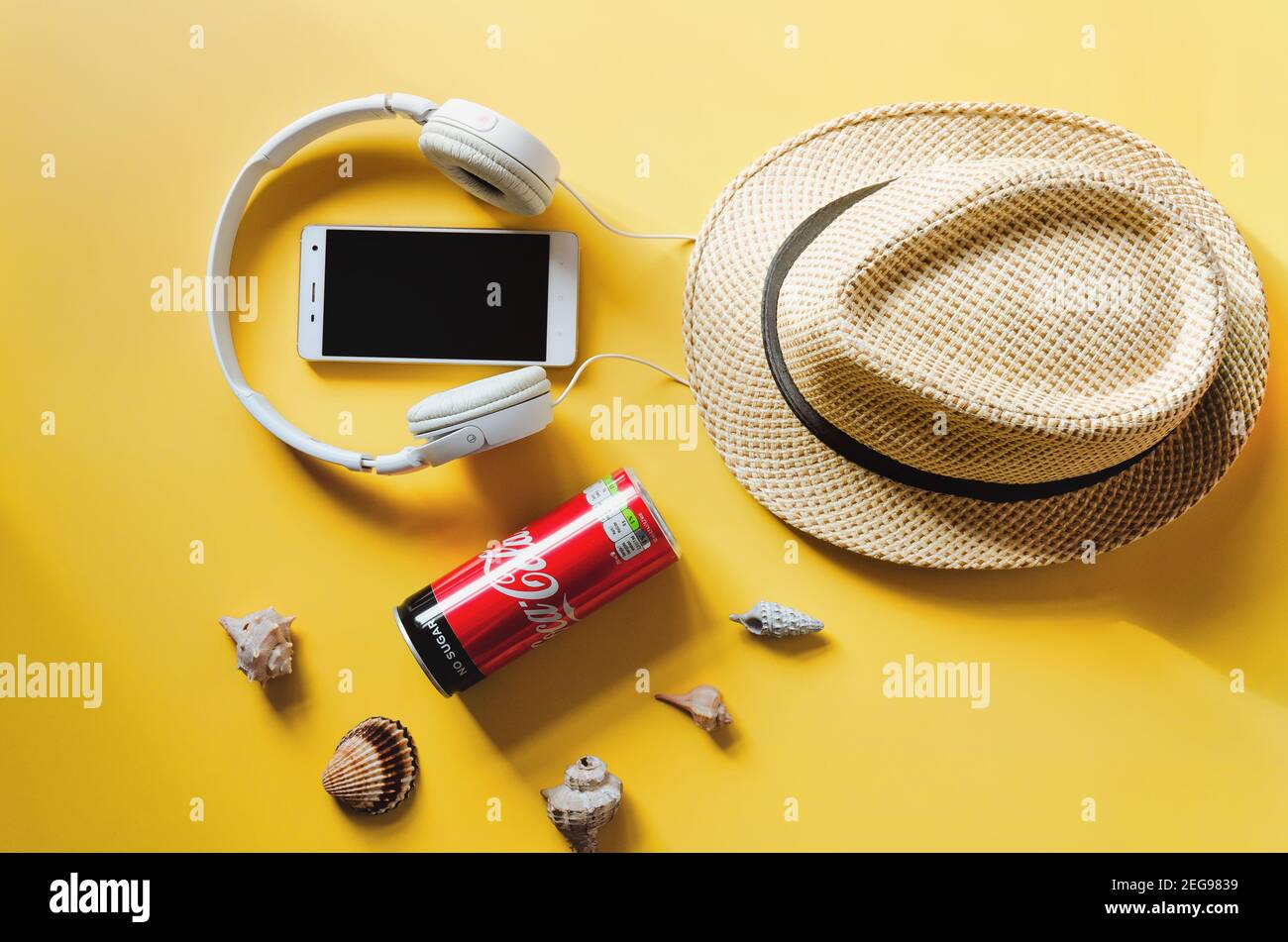 Travel under Covid-19 and new normal concepts. Top view of white headphones, can of Coca- Cola, phone and beach hat on yellow background close up Stock Photo