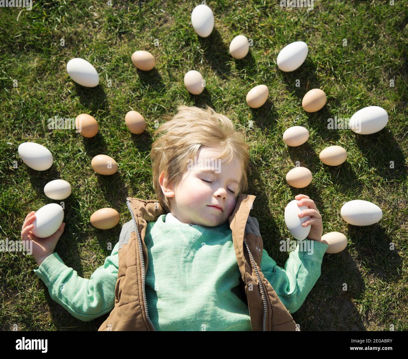 many white and beige chicken and goose eggs are laid out around the head of a 4-year-old blond boy sleeping on the grass. Cheerful preparation for Eas Stock Photo