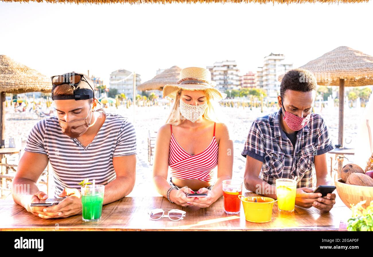 Multiracial friends with closed face masks using tracking app with mobile smartphones - Bored young milenial people at beach cocktail bar Stock Photo