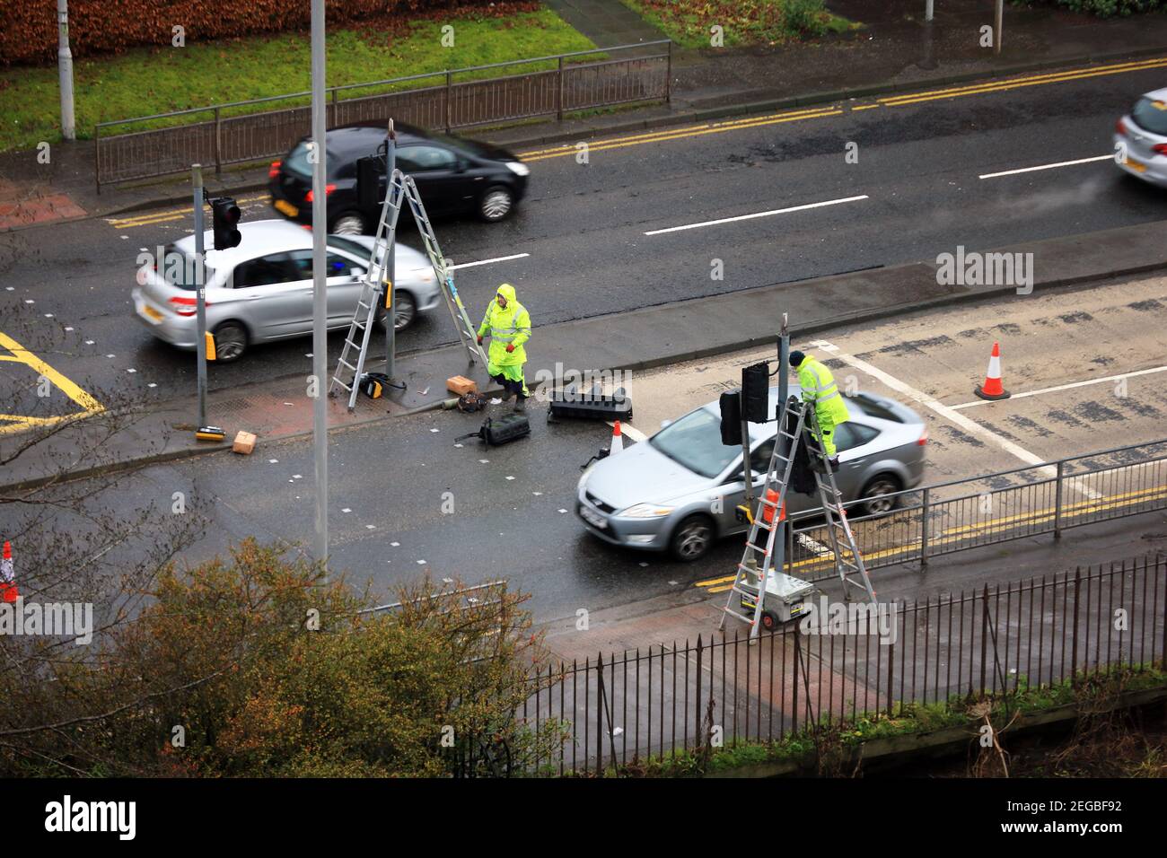 Workmen up ladders working on traffic lights, whilst cars pass, on a wet day in Paisley, Scotland Stock Photo