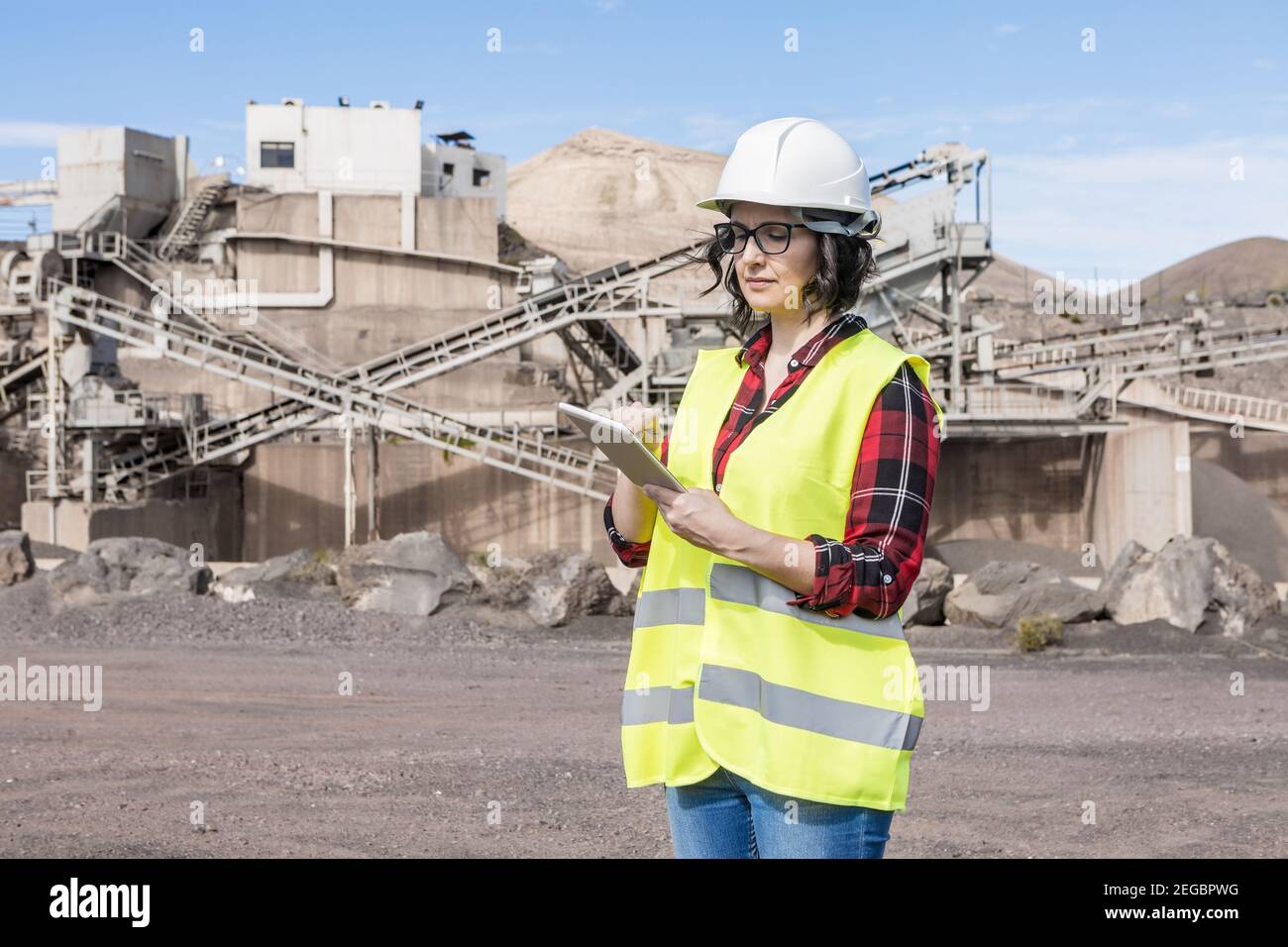 Professional female architect in hardhat and waistcoat checking draft on tablet while standing near industrial facility of construction site Stock Photo