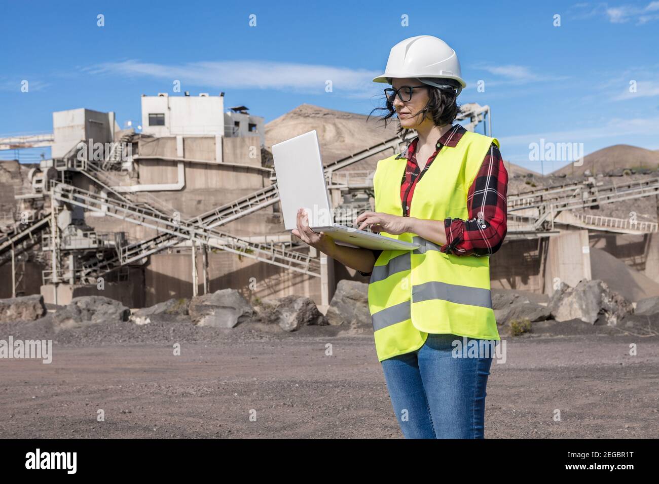 Serious female engineer in hardhat and waistcoat searching information on laptop while standing near industrial facility of construction site Stock Photo