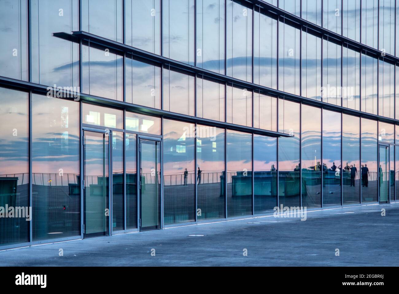 Sunset mirroring in facade of glass. The large glass facade reflects people and the evening sky. Sonnenuntergang spiegelt in großer Glasfassade. Stock Photo