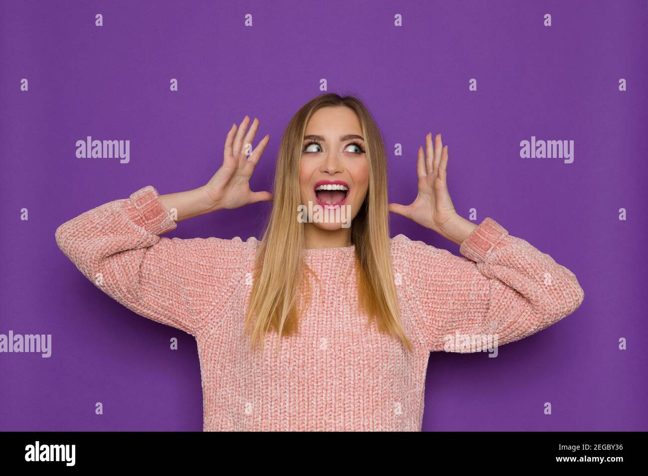 Happy young woman in pink sweater is holding head in hands, looking at the side and shouting. Front view. Waist up studio shot on purple background. Stock Photo