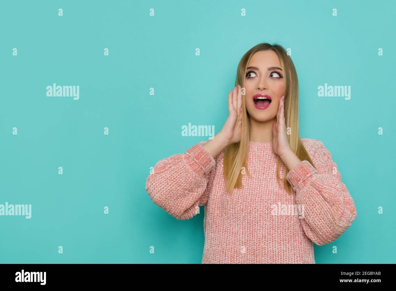 Surprised young woman in pink sweater is holding head in hands, looking at the side and talking. Front view. Waist up studio shot on turquoise backgro Stock Photo