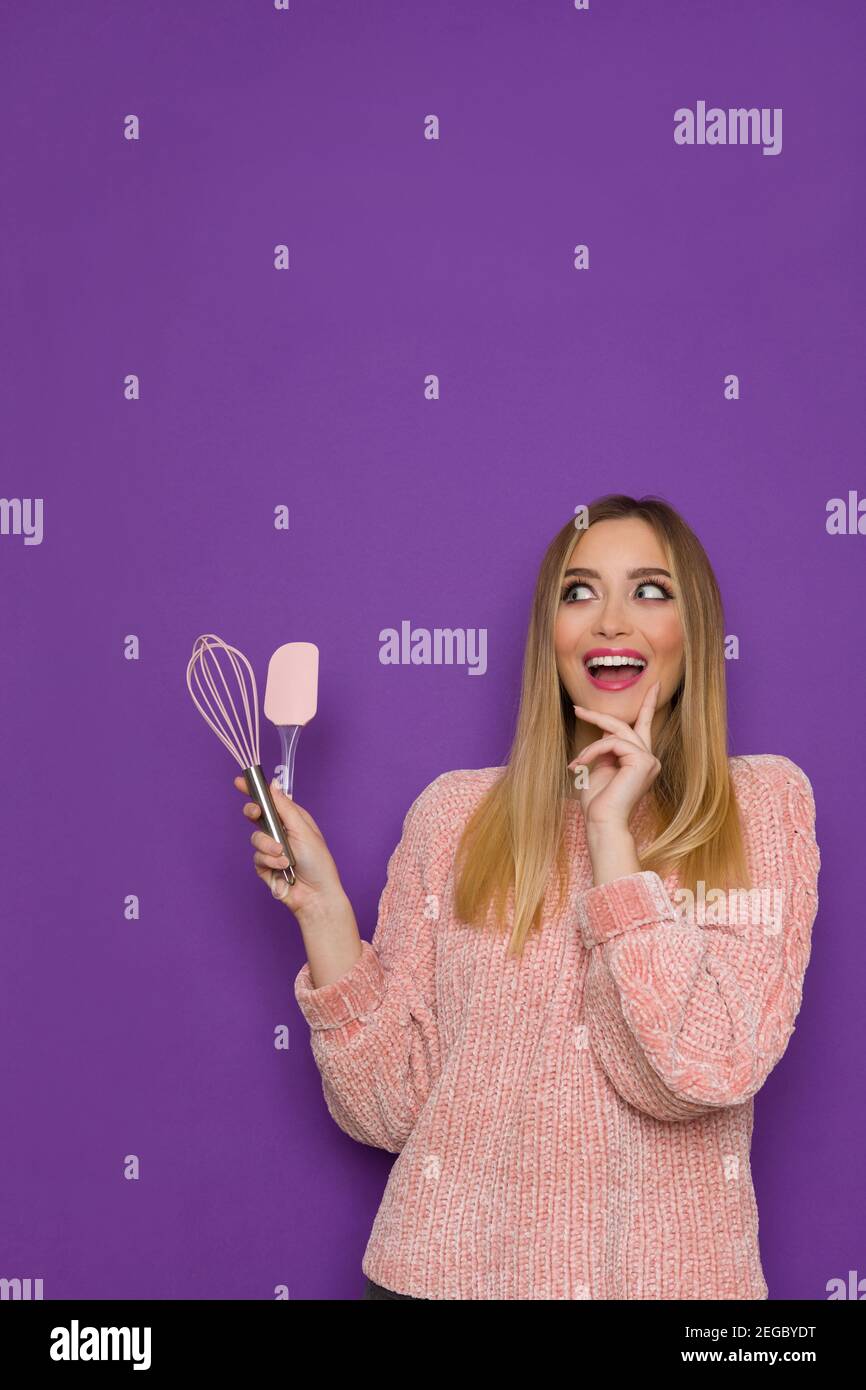 Happy young woman in pink sweater is holding kitchen tools, looking at the side and talking. Front view. Waist up studio shot on purple background. Stock Photo
