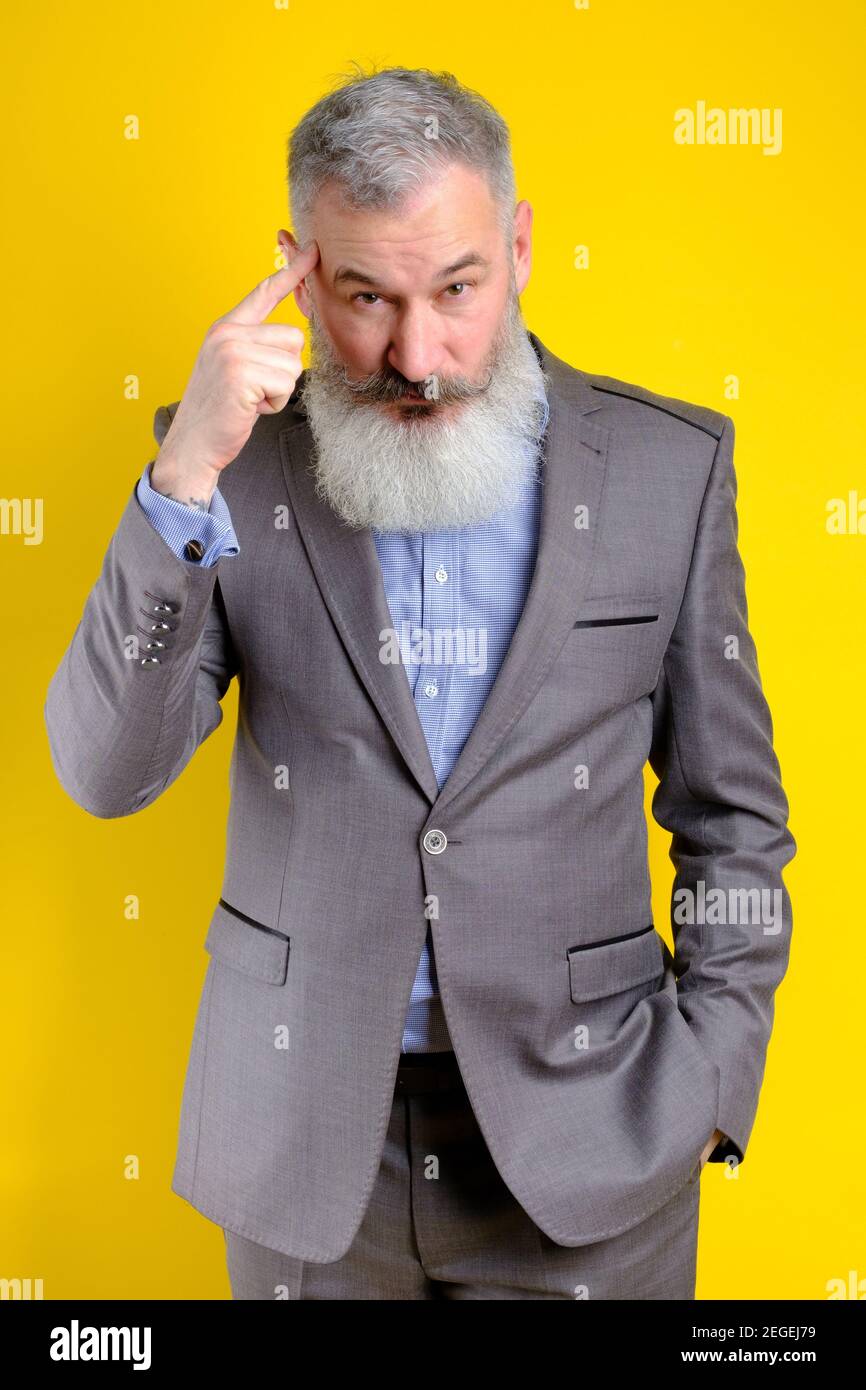Studio portrait mature businessman dressed in gray suit pointing to head with forefinger, great idea or thought concept, good memory, yellow backgroun Stock Photo