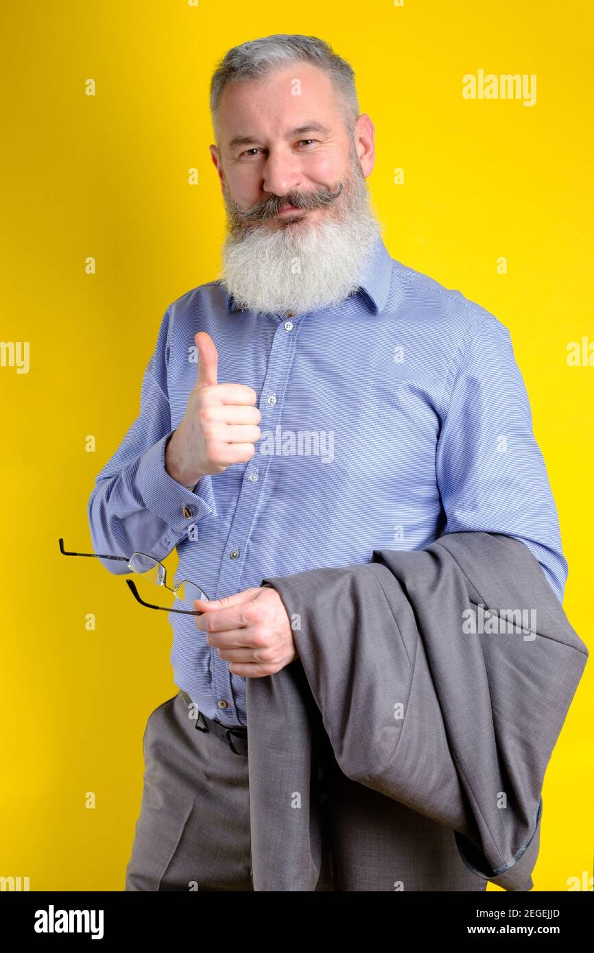 Studio portrait mature bearded businessman dressed in gray suit shows thumb up, successful business concept, yellow background Stock Photo